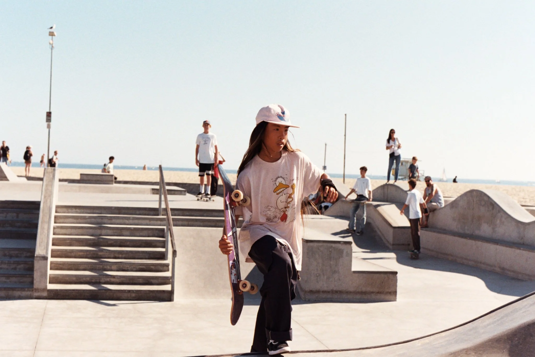 A young girl with long hair in a pink t-shirt, pants, and a white cap, practicing skateboard tricks at a skate park by the beach on a sunny day.