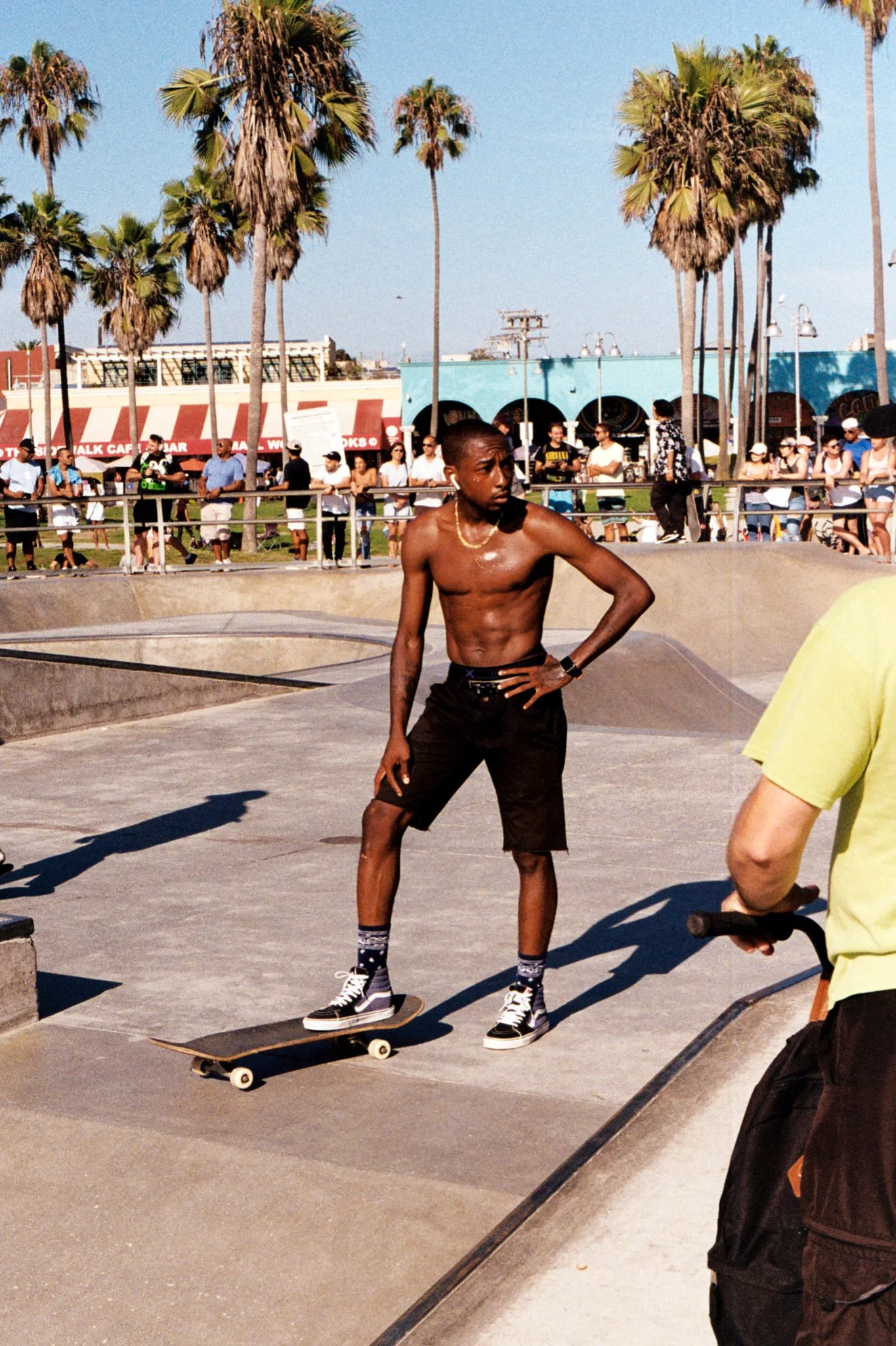 A shirtless young man with dark skin riding a skateboard at a skatepark, surrounded by people and palm trees under a clear blue sky.
