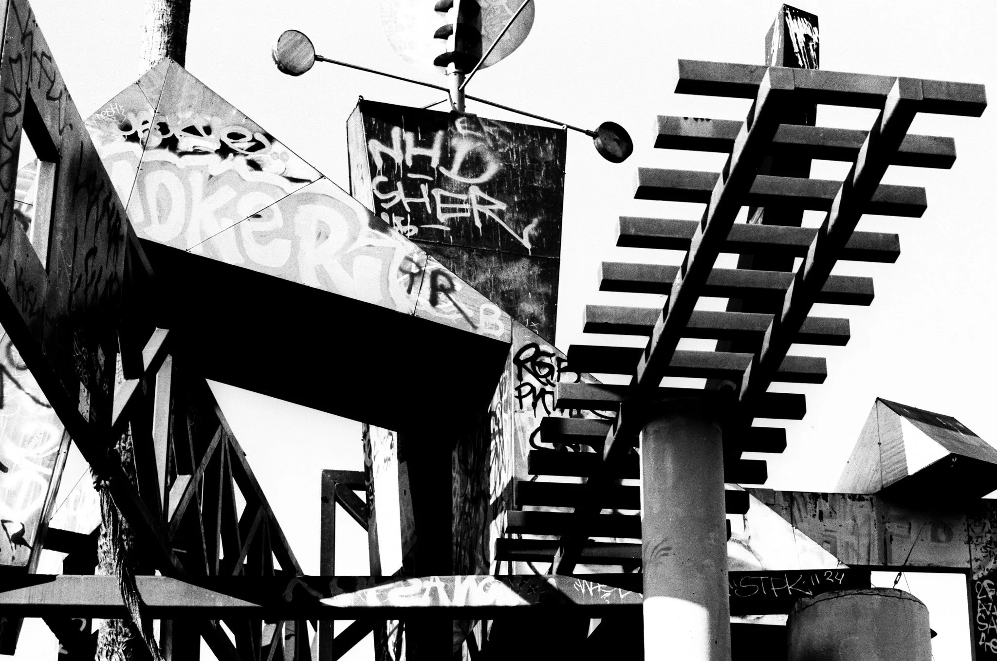 Black and white photograph of urban structures with graffiti, including metal beams, stairs, and signage.