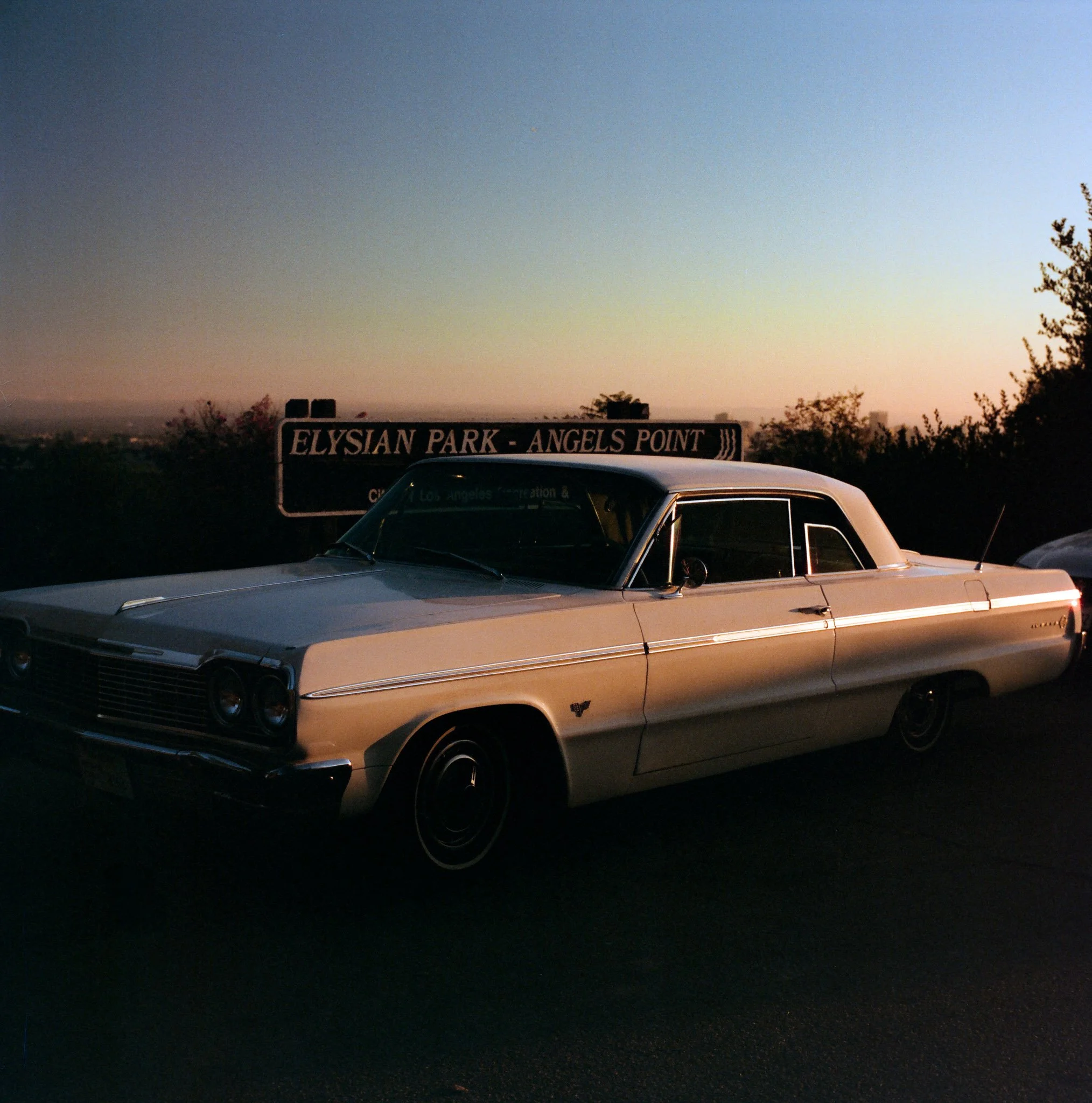 A vintage cream-colored Cadillac parked at Elysian Park, Angels Point, during sunset with a sunset sky in the background and trees surrounding the area.