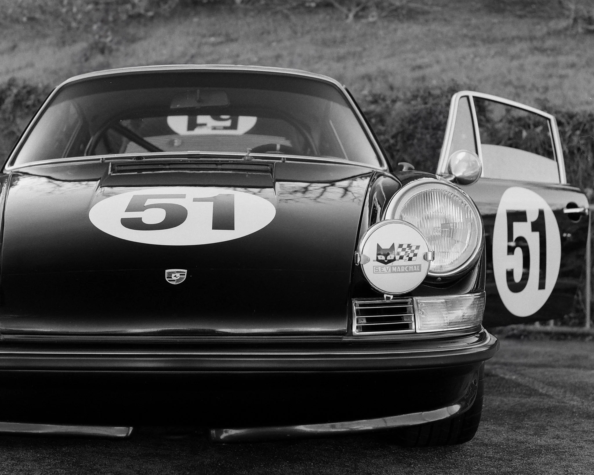 A black and white photograph of a vintage racing car with the number 51 on its hood and door, parked outdoors with a sponsor badge and a racing event sign in the background.