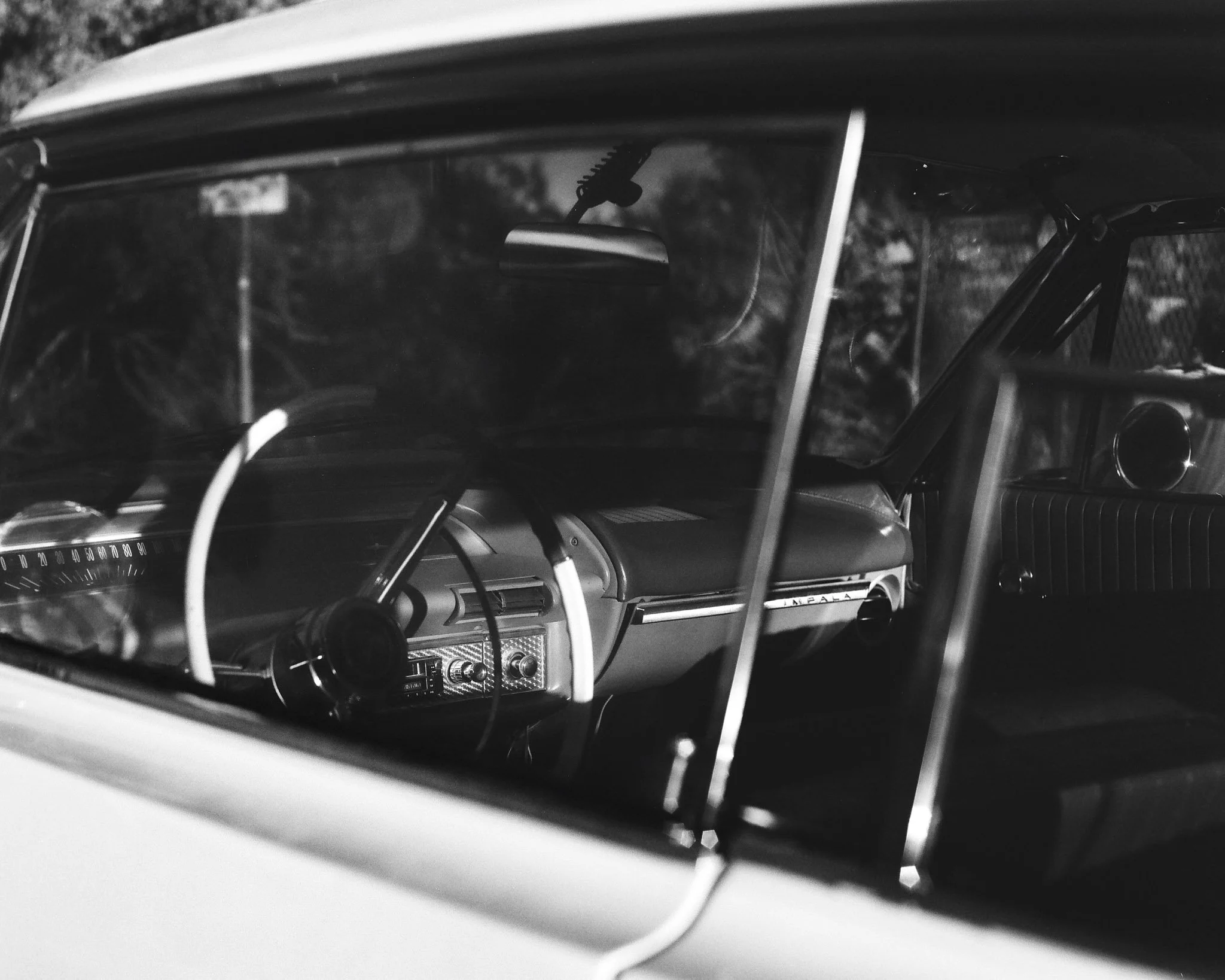 Black-and-white photo of the interior of a vintage car viewed through the front windshield, showing the steering wheel, dashboard, and driver’s seat.