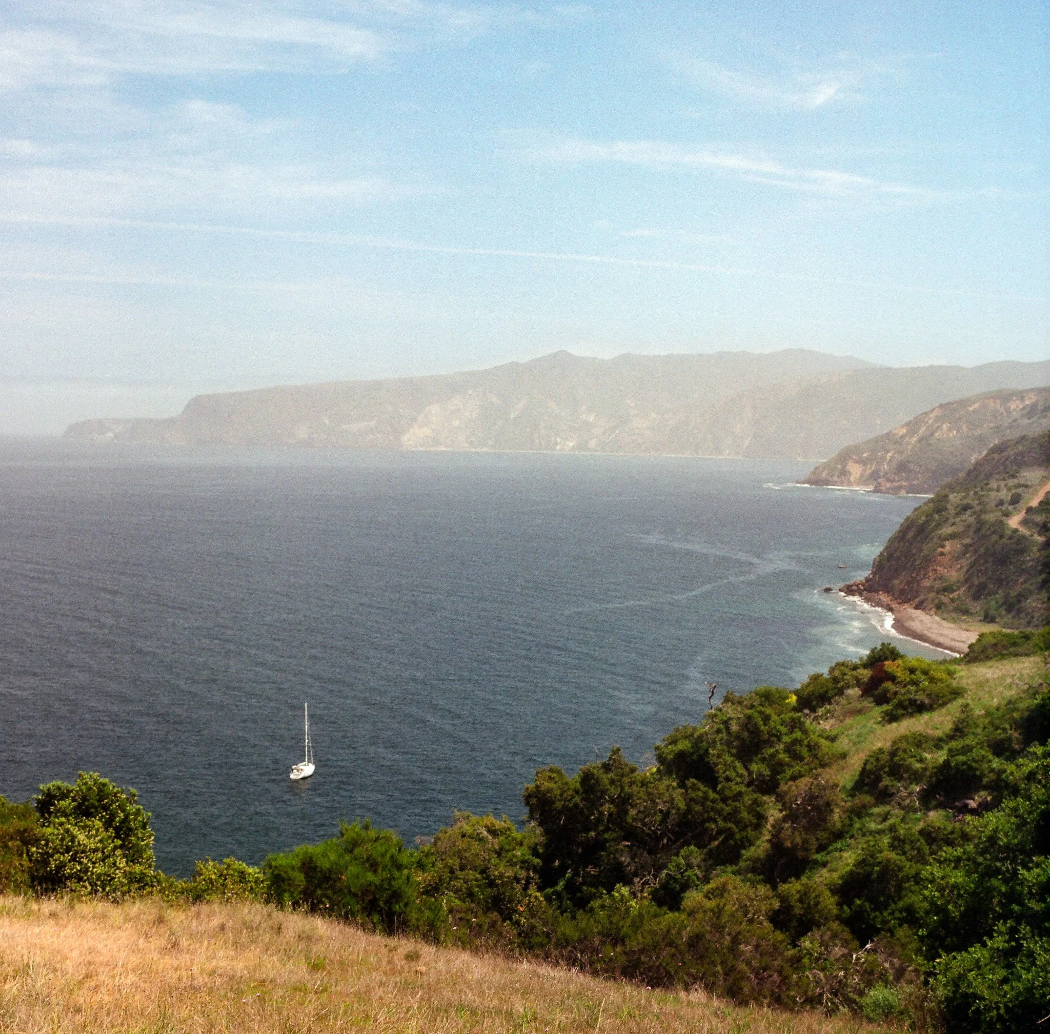View of the ocean with a sailboat, green hills, and distant mountains under a blue sky with wispy clouds.