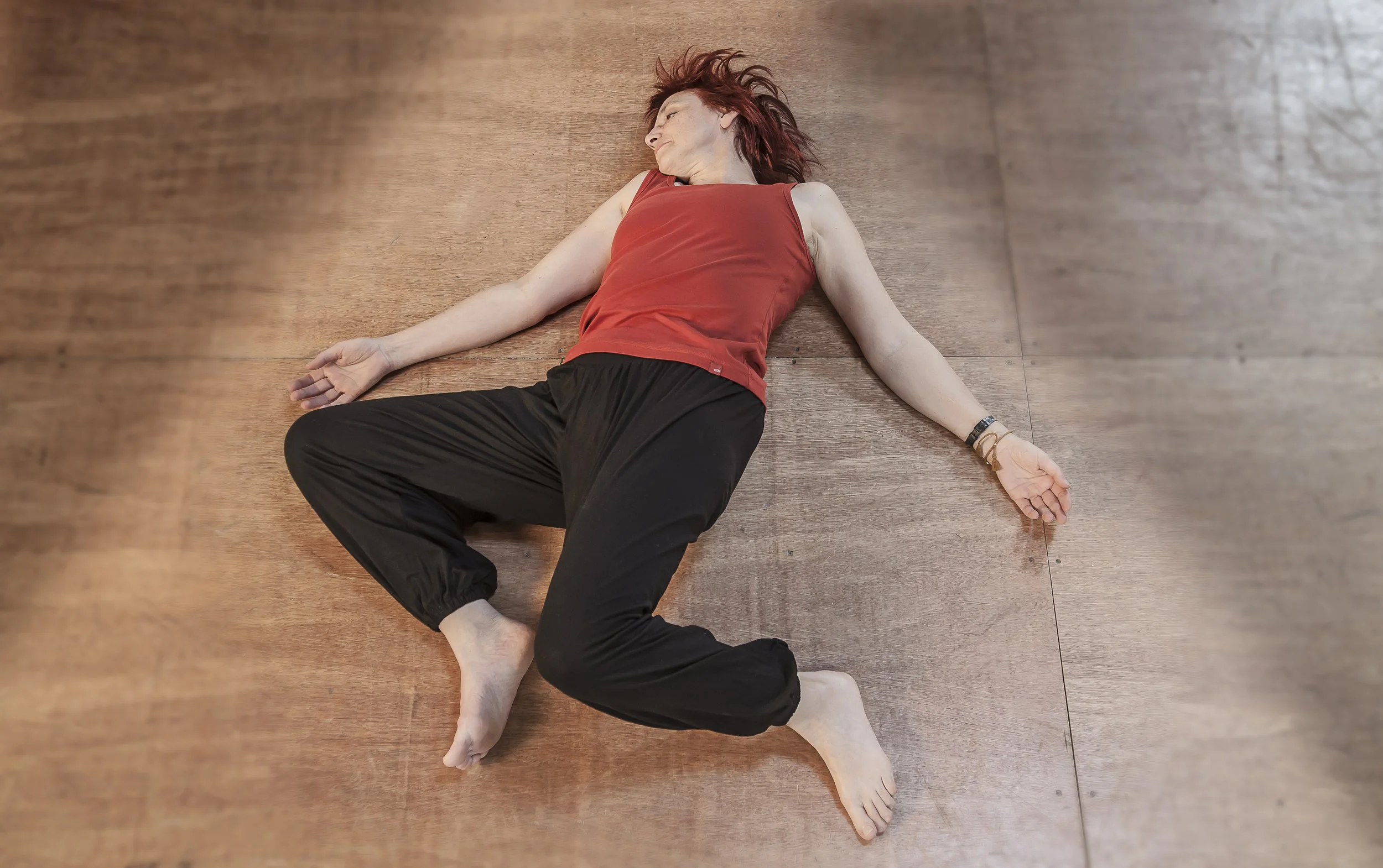A woman lying on the floor with arms stretched and knees bent during an Awareness Through Movement® lesson while listening to themselves within movement directions given by a teacher.