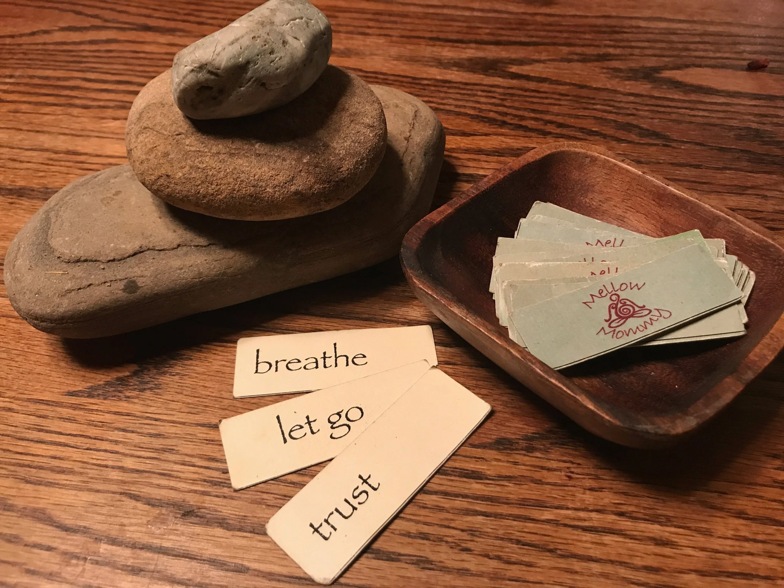 Stack of four smooth stones on a wooden surface, with a small wooden bowl containing printed cards that say "Mellow Momma" and a few paper slips with words "breathe," "let go," and "trust" on them.