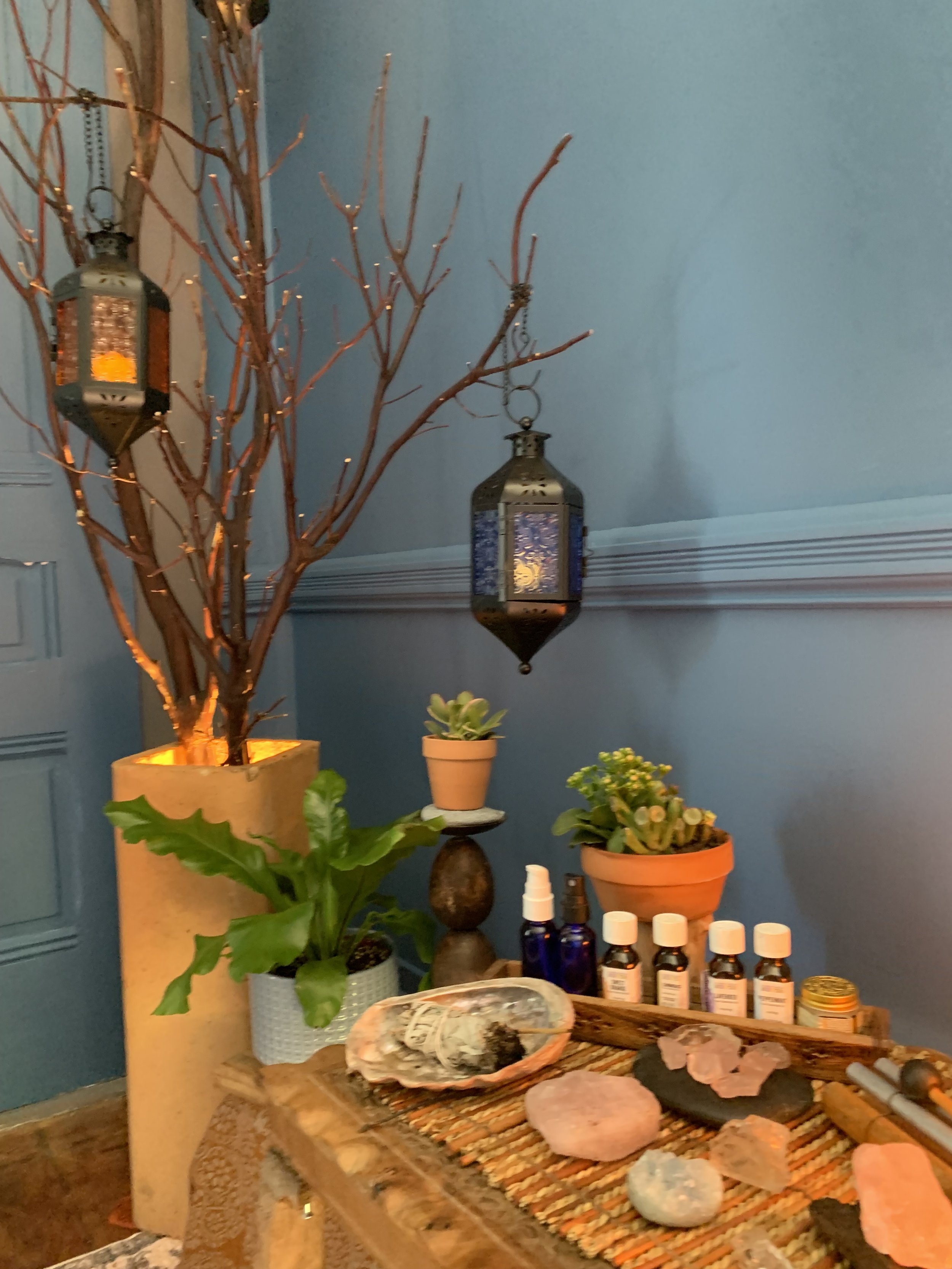 Cozy room decor with lanterns, potted plants, scented oils, and crystals on a wooden table.