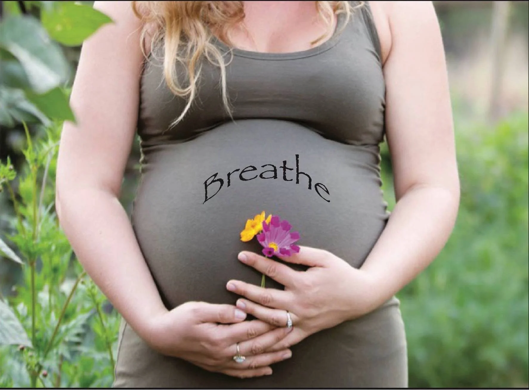 Pregnant woman in a green tank top holding pink and yellow flowers over her belly with the word 'breathe' written on it, standing outdoors with greenery in the background.
