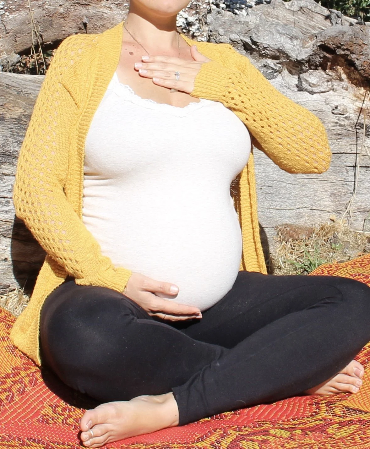 A pregnant woman sitting cross-legged outdoors on a red woven blanket, wearing a white top and black pants, with a yellow cardigan, with her left hand on her belly and her right hand on her chest.