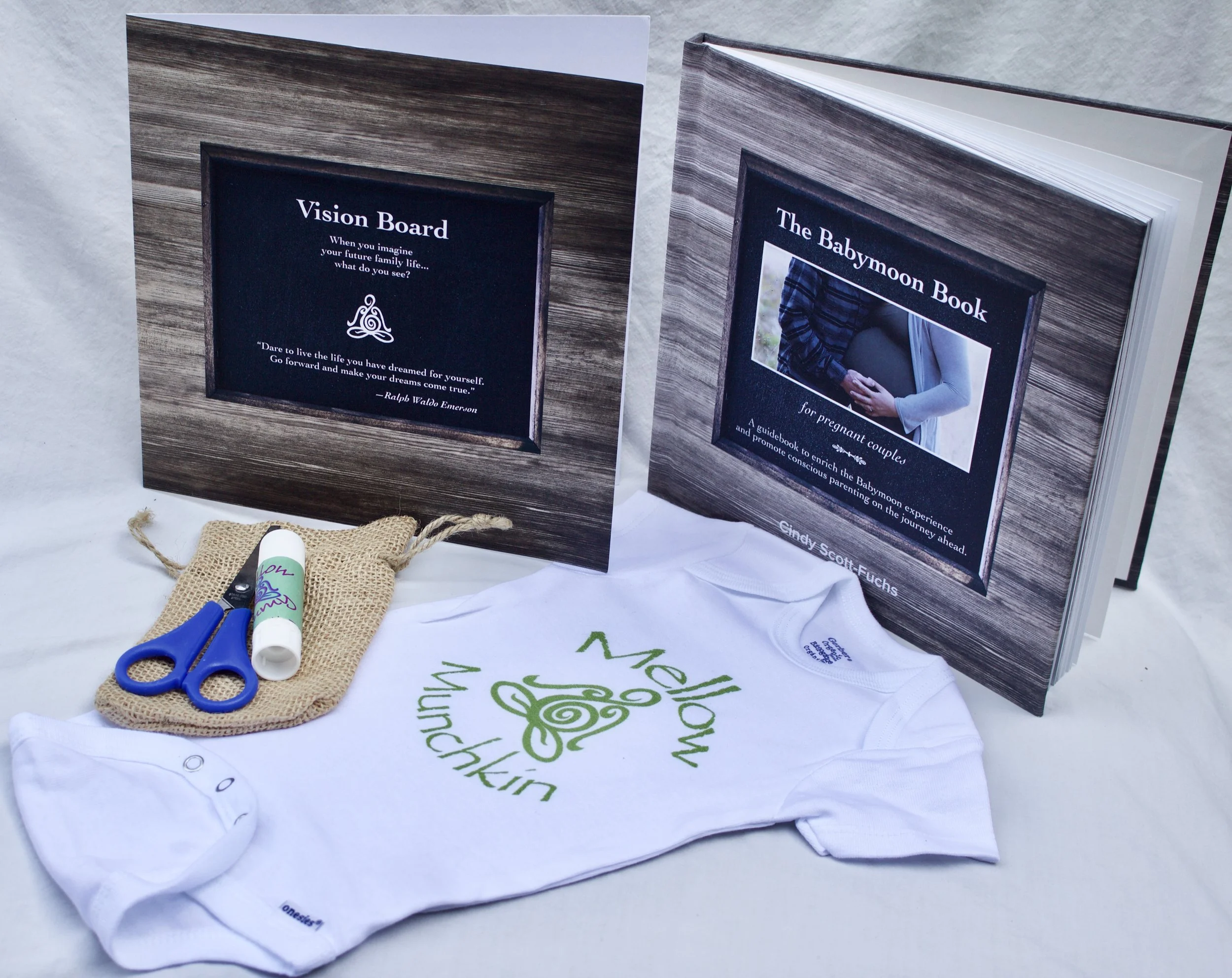 An arrangement of pregnancy planning items including two open books, a white onesie, a pair of blue scissors, a small burlap pouch, and a white tube of lotion. The books are titled 'Vision Board' and 'The Babymoon Book.'