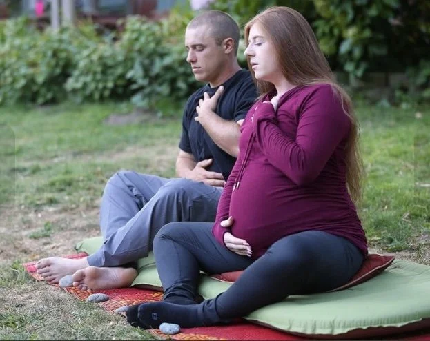A pregnant woman sitting on a cushion outdoors with a man, both with eyes closed, holding their chests and belly, appearing to meditate or relax.