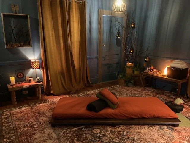 A dimly lit room with a bed on the floor, covered with an orange blanket and pillows. There are candles, lamps, and decorative items on side tables, with curtains and a door in the background. The room has a cozy, relaxed atmosphere.