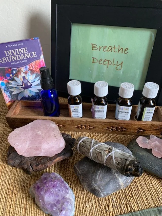 A collection of mindfulness and spiritual items, including a framed print reading "Breathe Deeply," essential oil bottles labeled with scents like lavender and peppermint, a bundle of sage, crystals including rose quartz and amethyst, a blue spray bottle, and a deck of Divine Abundance oracle cards.