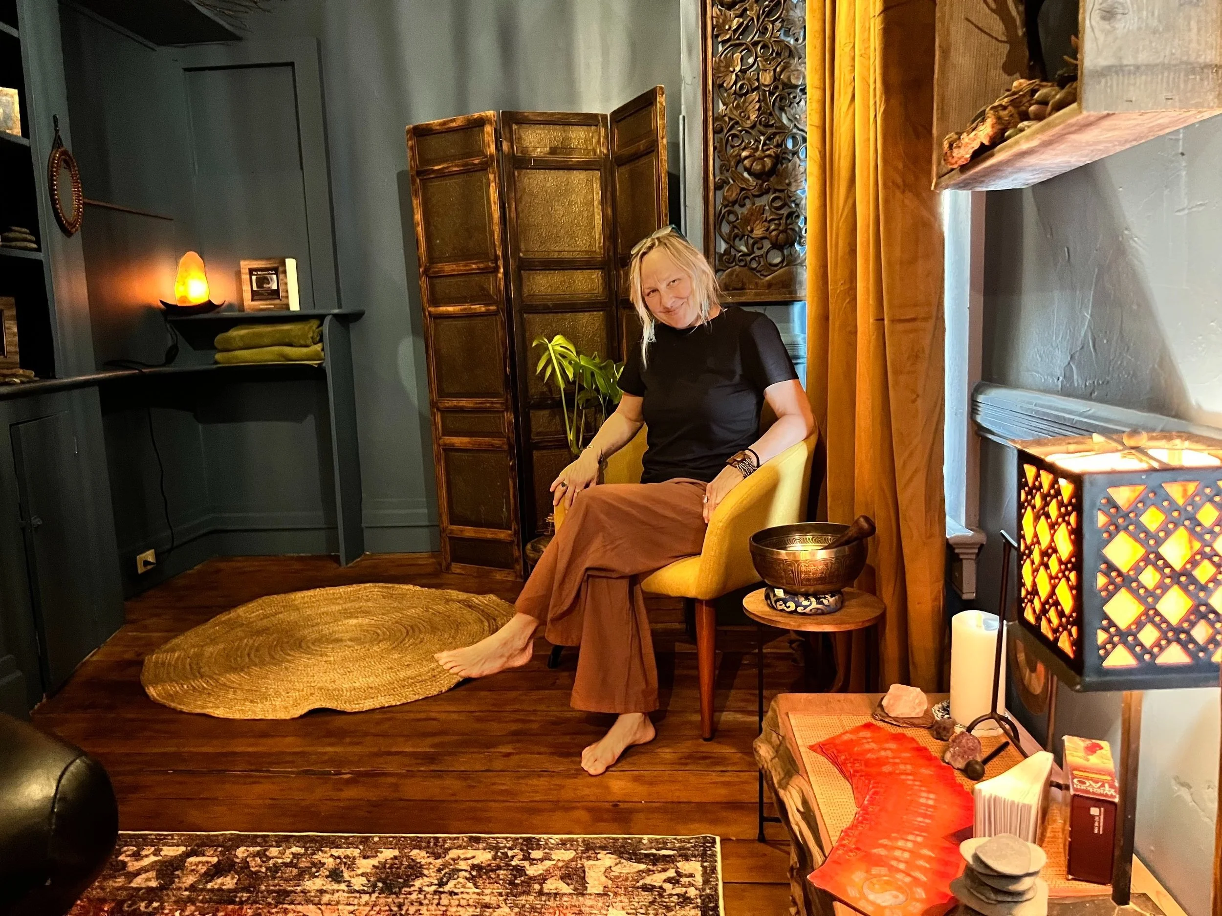 A woman sitting in a cozy, dimly lit room with wooden floors and blue walls. She is seated in a yellow chair next to a small table with a decorative item. The room features a rug, a decorative lamp with a glowing design, and various items such as a Himalayan salt lamp and stacked towels. A wooden screen and wall art are in the background, contributing to a relaxing atmosphere.