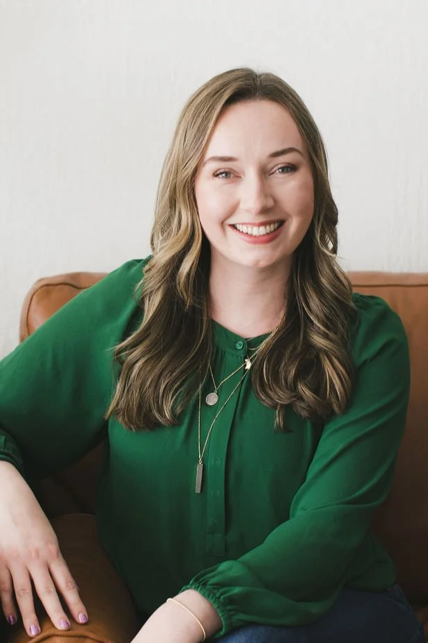 A woman with long wavy hair wearing a green blouse, sitting on a brown leather chair, smiling at the camera.