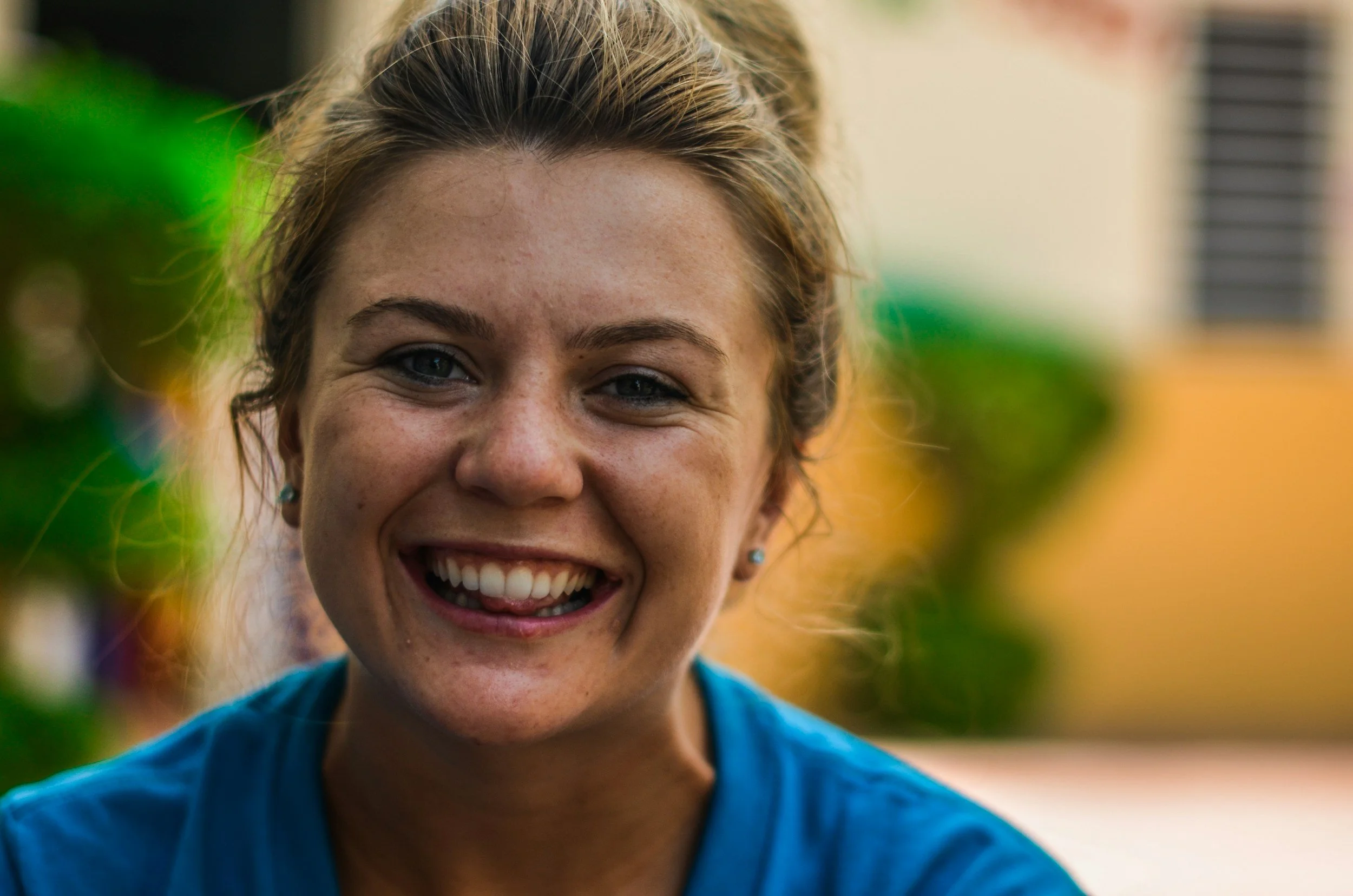 Close-up of a smiling woman with brown hair, blue eyes, freckles, and earrings, wearing a blue shirt, outdoors with blurred greenery and a building in the background.