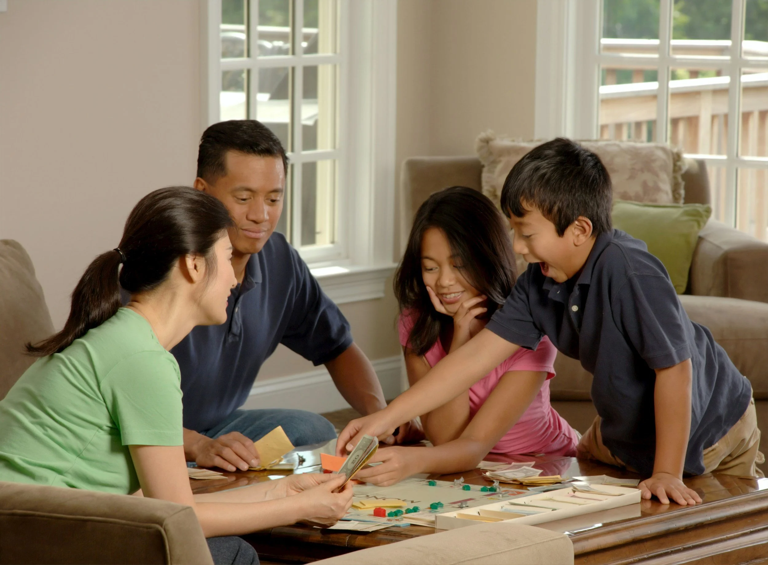 Family playing Monopoly game around the table in living room, with sunlight coming through large windows.