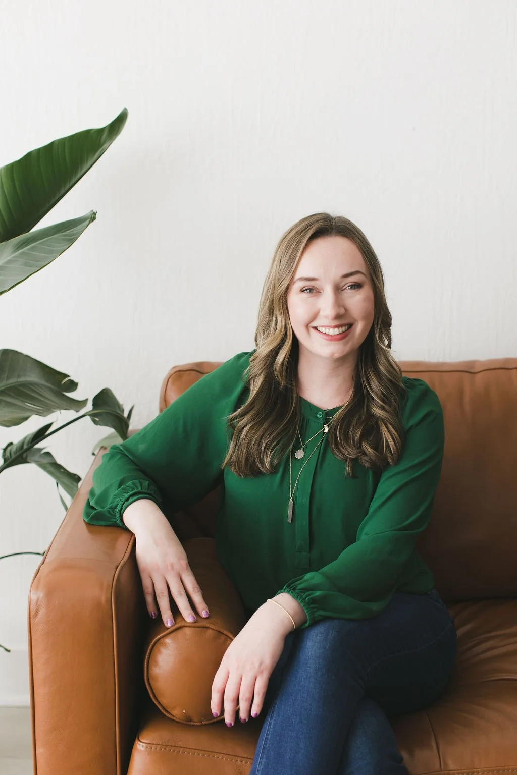 A smiling woman with wavy brown hair, wearing a green blouse and blue jeans, sitting on a brown leather sofa next to a large green houseplant against a white wall.