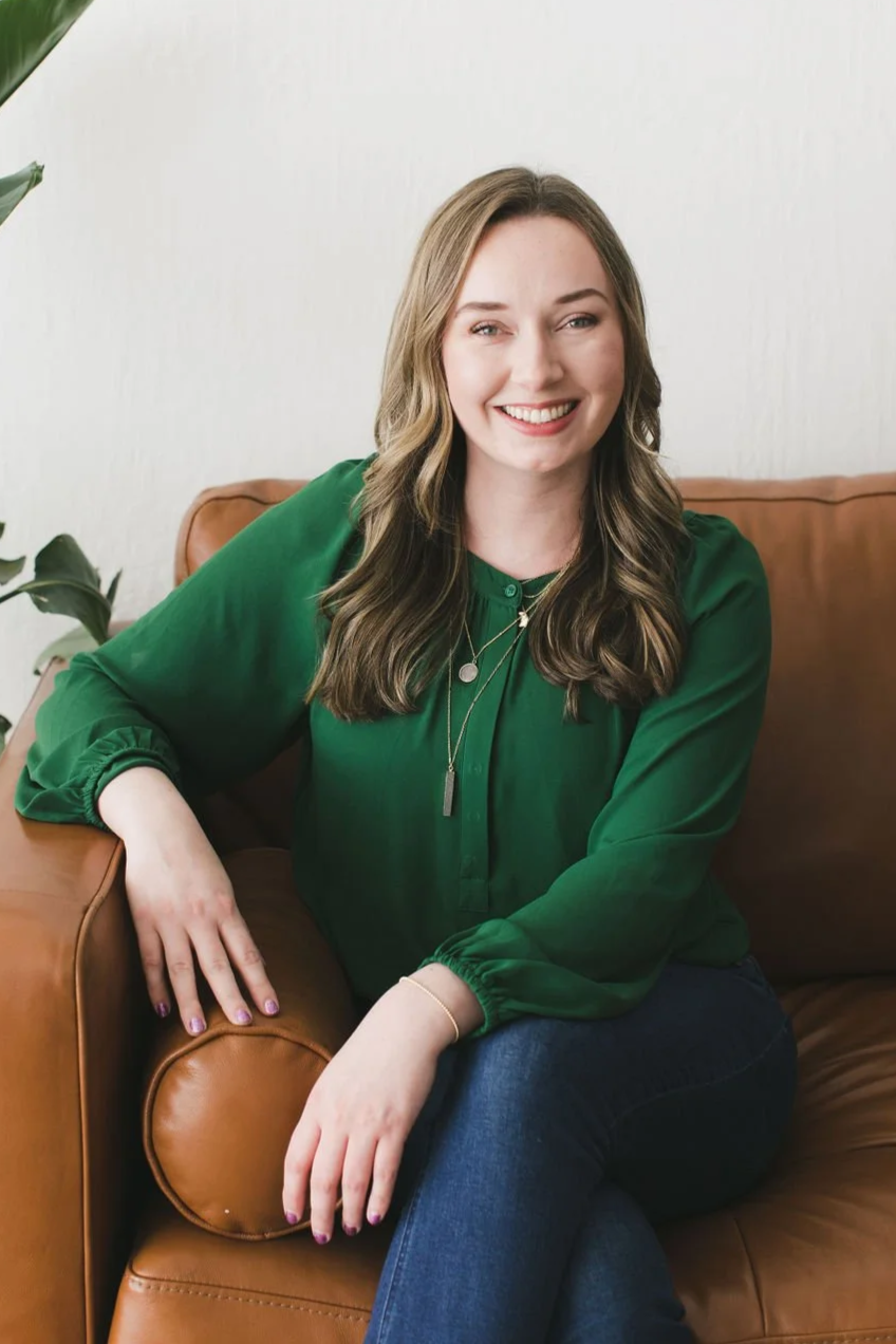 A smiling woman with wavy brown hair wearing a green blouse and dark jeans, sitting on a tan leather sofa next to a large green plant against a white wall.