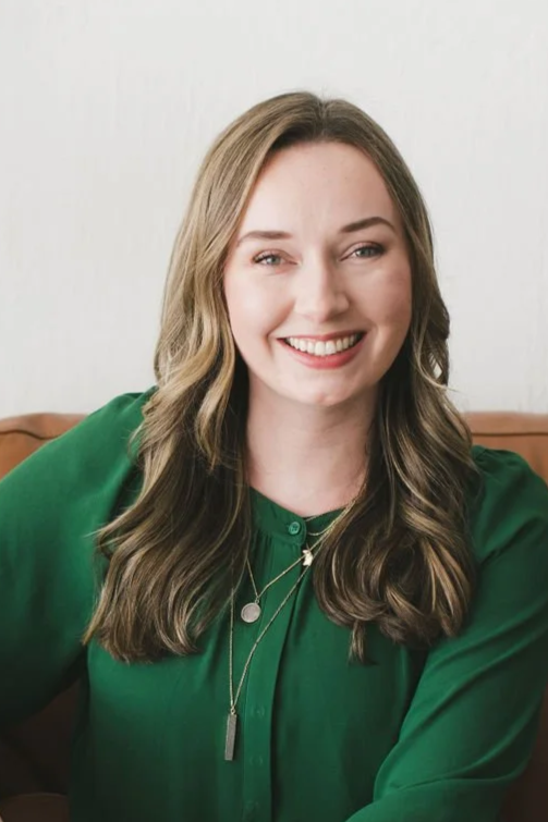 A smiling woman with wavy brown hair wearing a green blouse and dark jeans, sitting on a tan leather sofa next to a large green plant against a white wall.