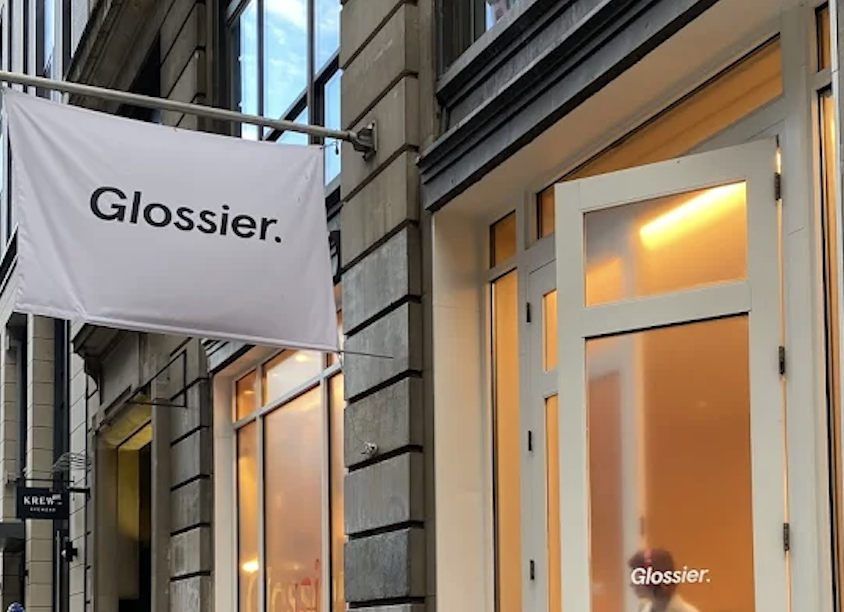 Exterior view of a building with large windows and a white door, with a white flag displaying the word 'Glossier.' hanging in front.