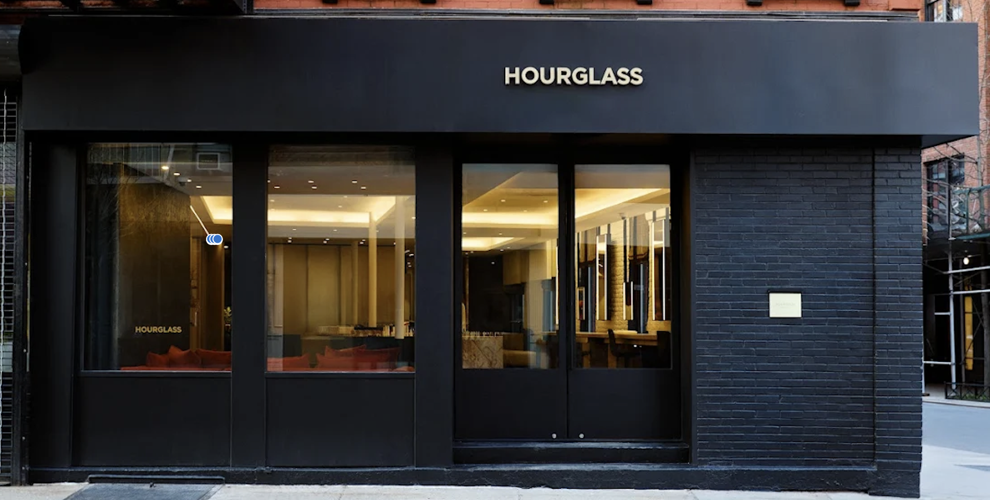 Exterior view of a modern black brick storefront with large glass windows and door, displaying the name 'HOURGLASS' above the entrance, illuminated interior lighting visible through the windows.
