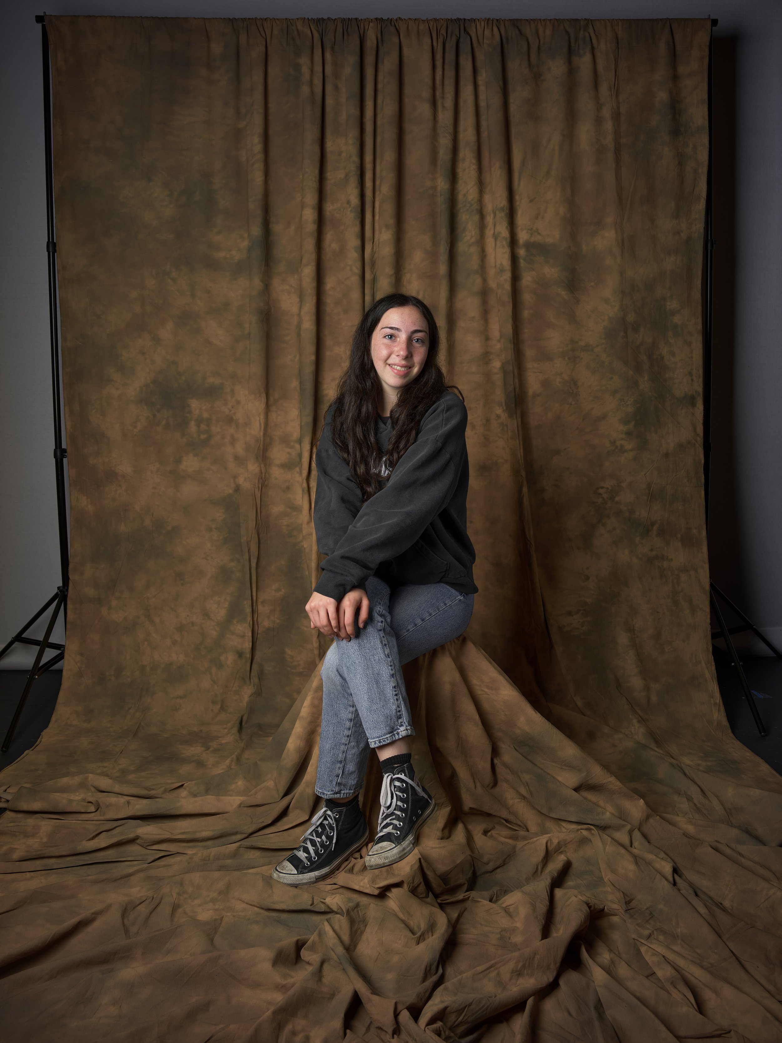 A young woman with long dark hair, wearing a black sweatshirt, jeans, and black sneakers, sits on a stool in front of a brown and green textured backdrop, smiling at the camera.