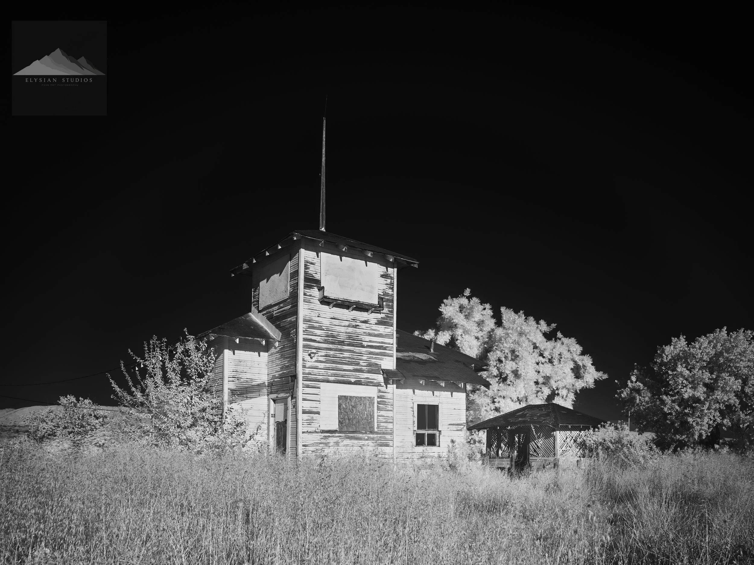 Black and white photo of an old, weathered wooden house with a steeple, surrounded by grass and trees, under a clear sky.