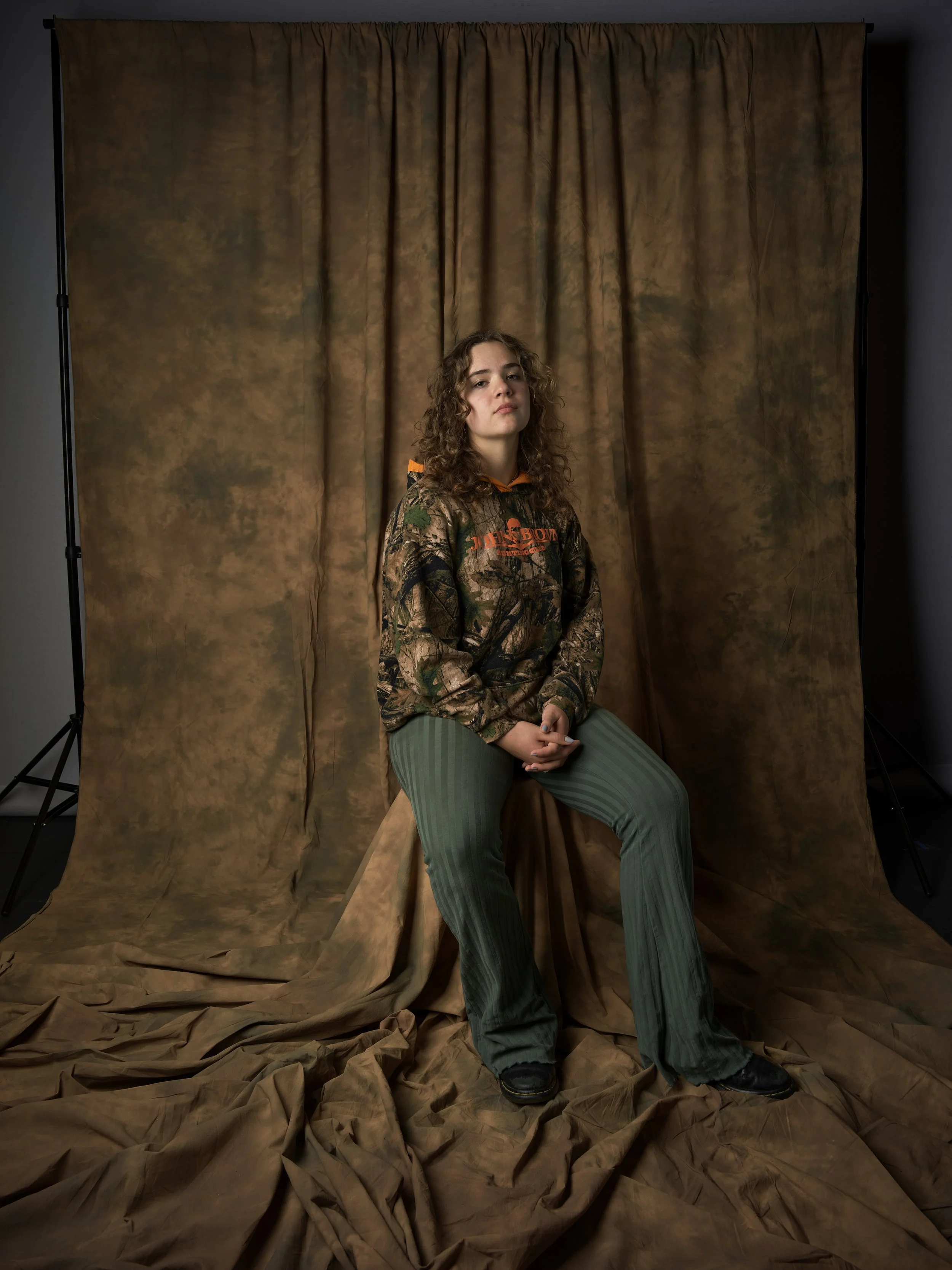 Young woman with curly hair sitting on a wooden stool in front of a brown backdrop, wearing a camouflage hoodie and green pants, with a serious expression.