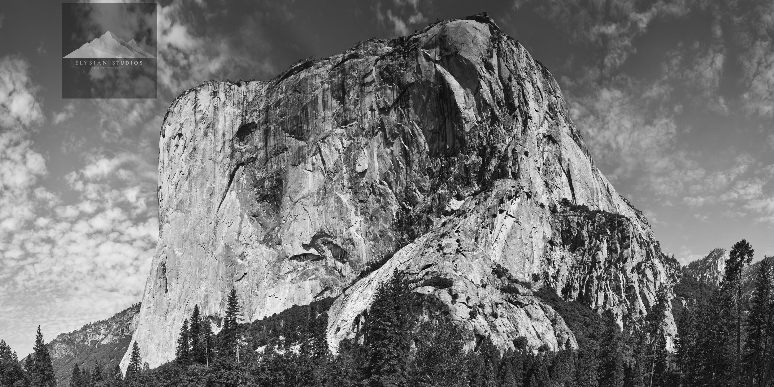 Black and white photo of a large mountain with a sheer, smooth face, surrounded by trees at its base and a cloudy sky above.