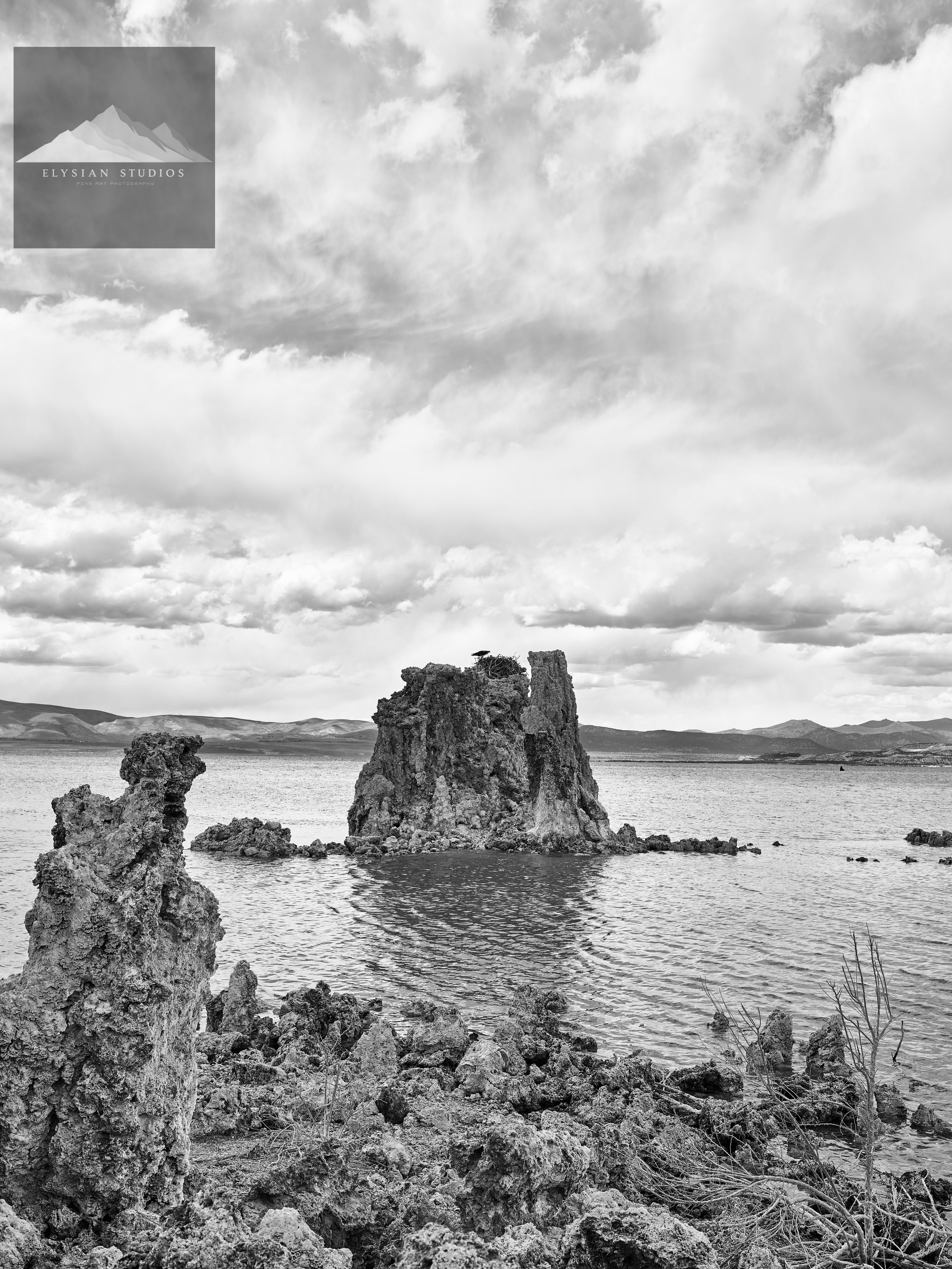 Black and white photo of large rock formations on the shoreline of a lake or river, with distant mountains and a cloudy sky in the background.