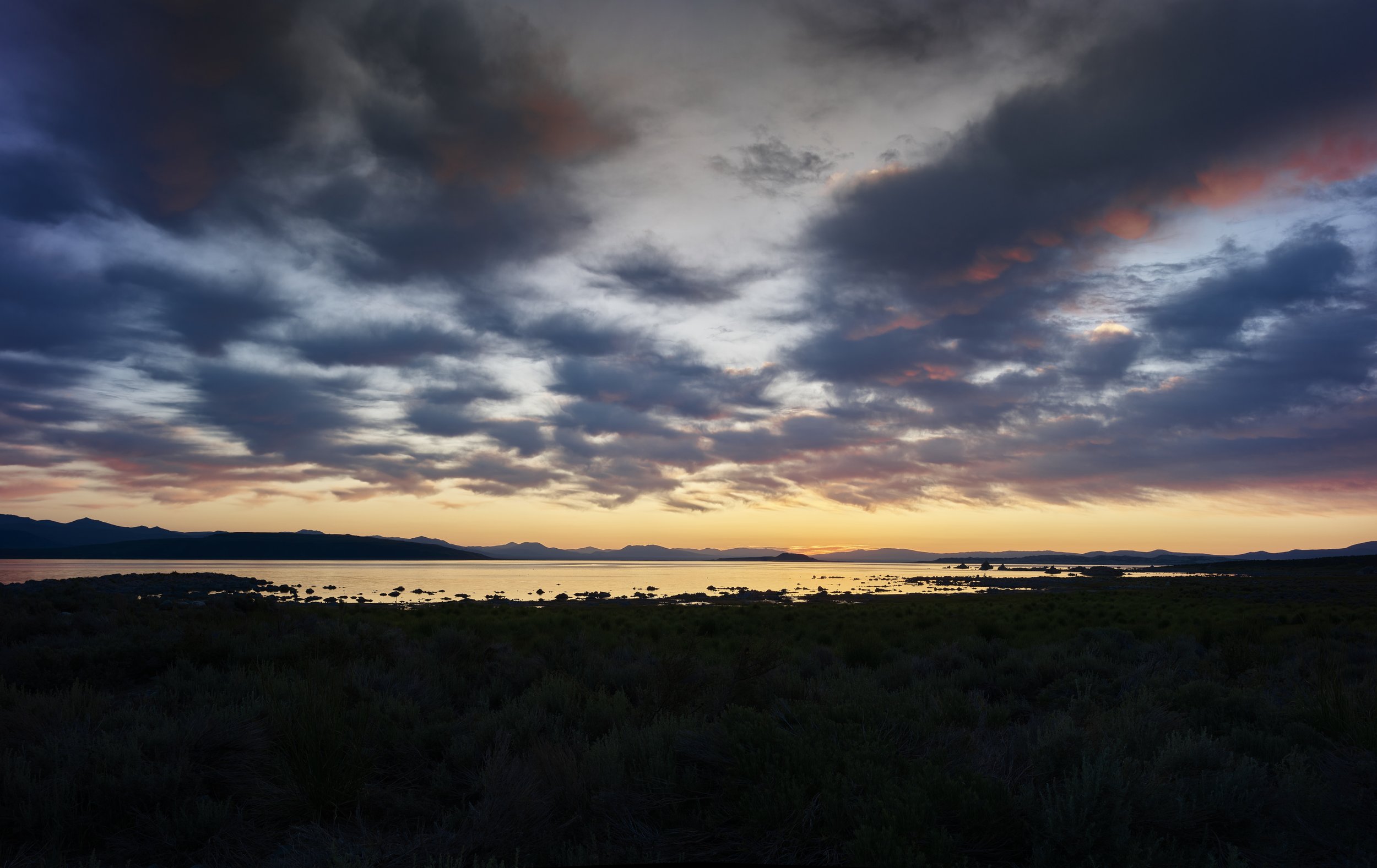 Sunset over a calm body of water with mountains in the background and cloudy sky with hints of pink and orange.