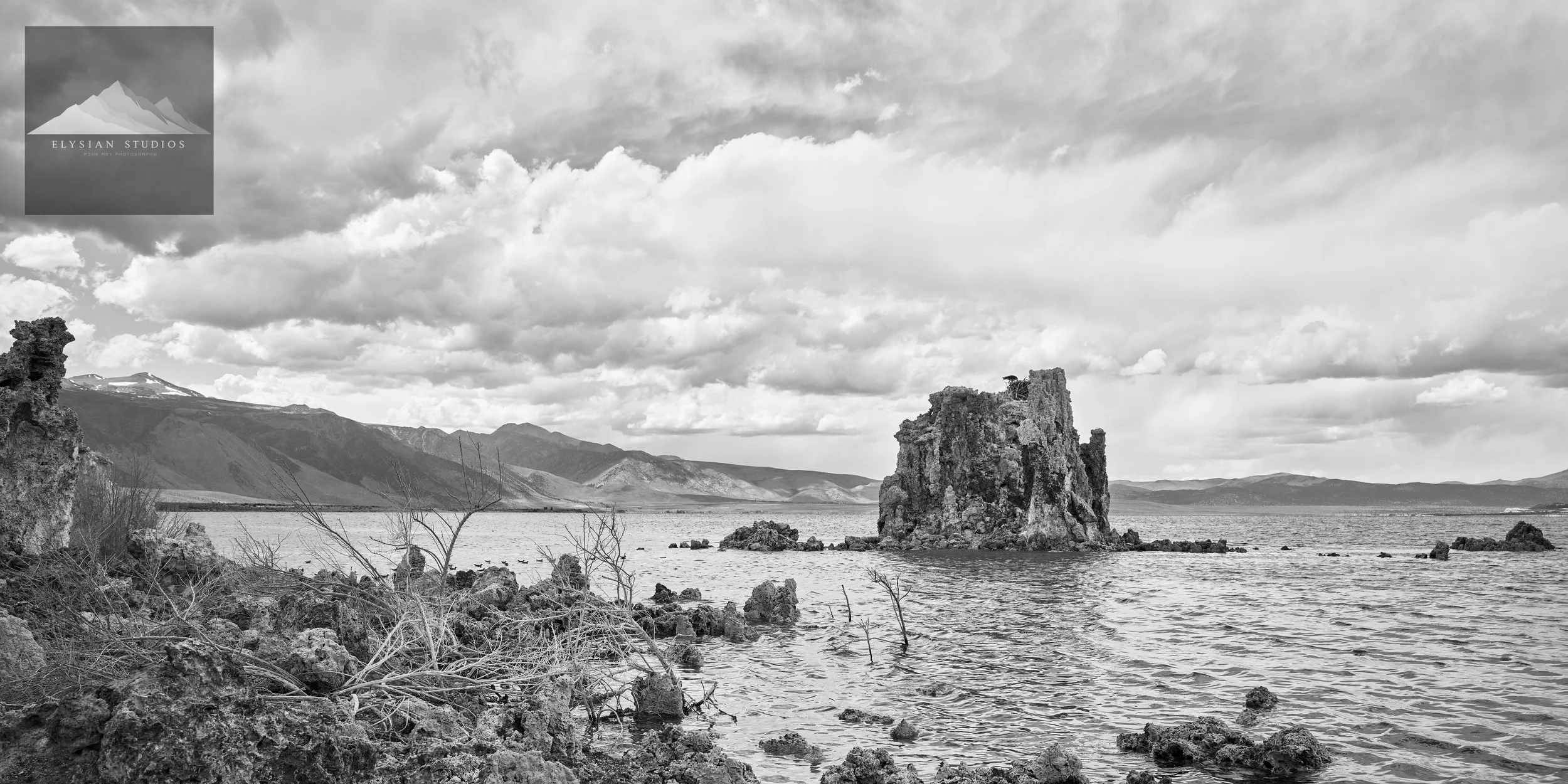 A black and white photograph of a rocky shoreline with a large rock formation rising from the water, mountain range in the background, and a cloudy sky overhead.
