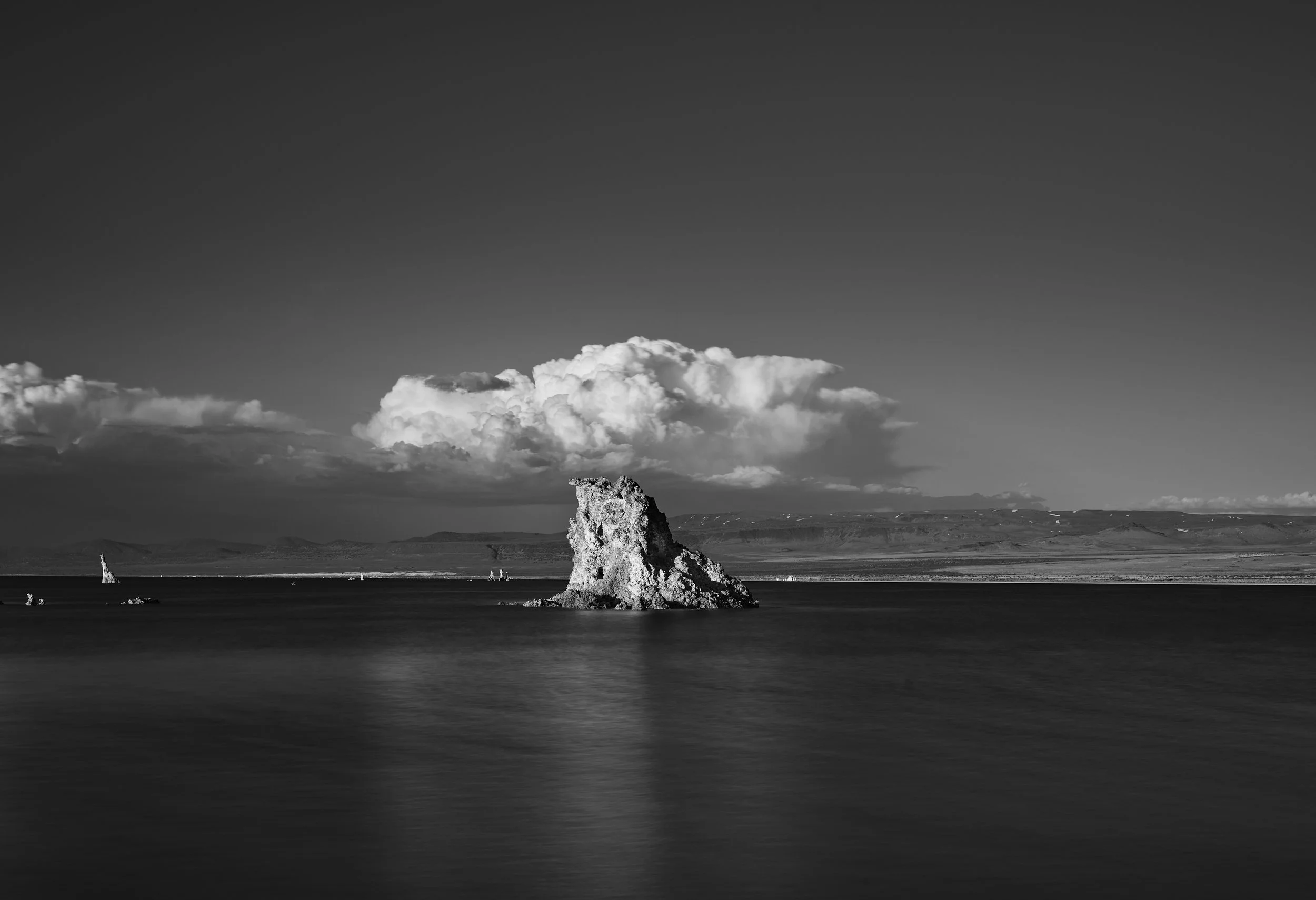 Black and white landscape photo of a large rock formation in the water with clouds in the sky and distant hills.