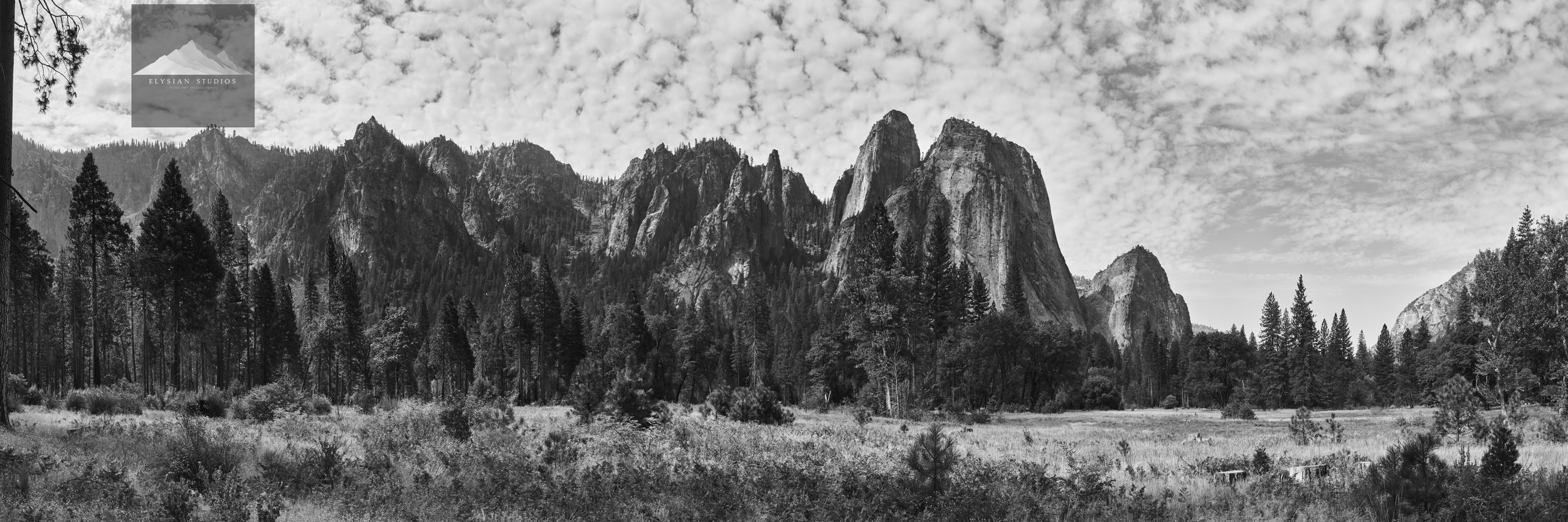 Black and white panoramic view of a mountainous landscape with tall, rugged cliffs and forested area at the base, under a sky filled with scattered clouds.