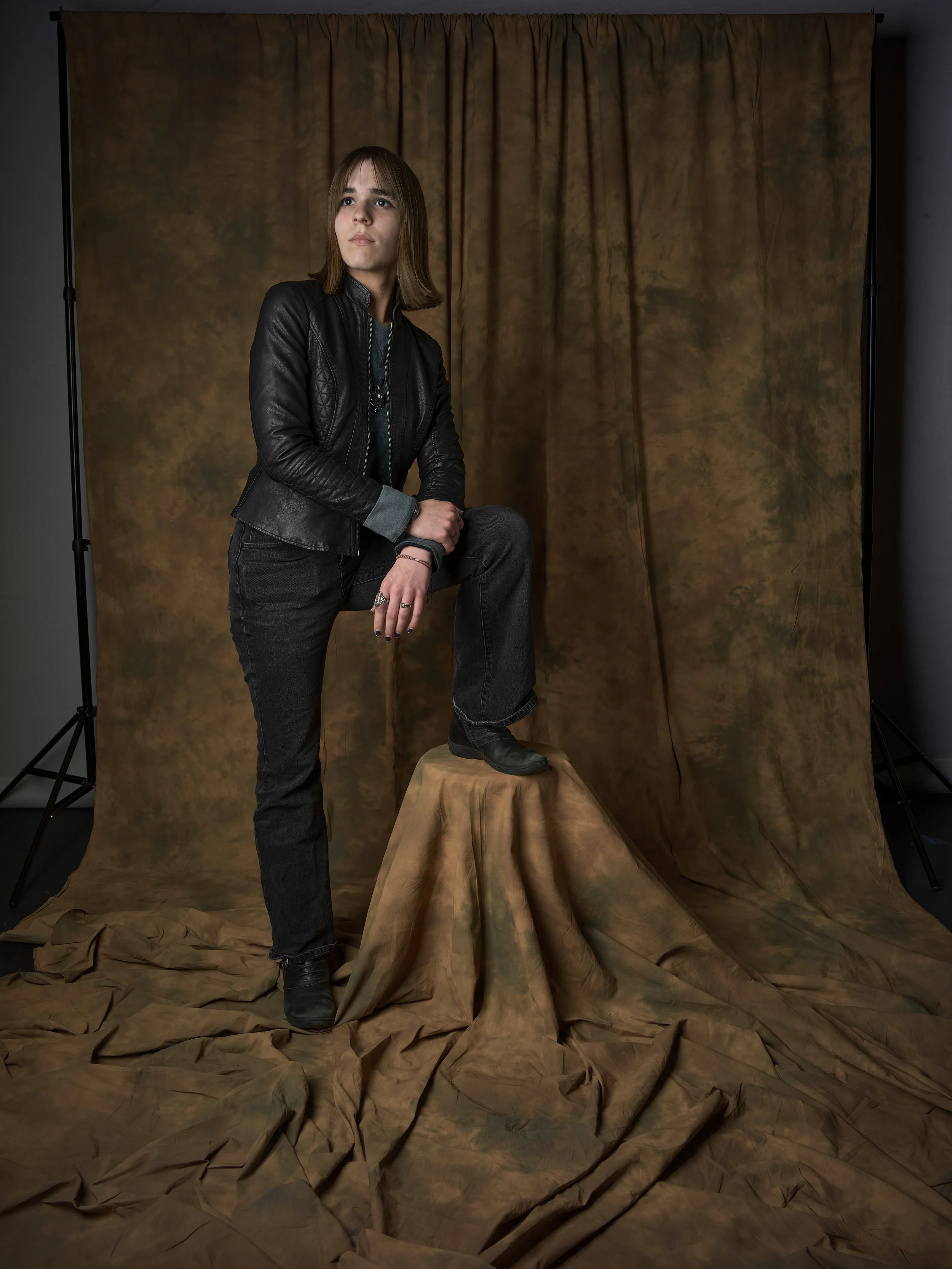 A young person with shoulder-length hair wearing a black leather jacket, black jeans, and black boots, standing with one foot on a draped fabric-covered pedestal, posed against a brown textured backdrop in a photography studio.