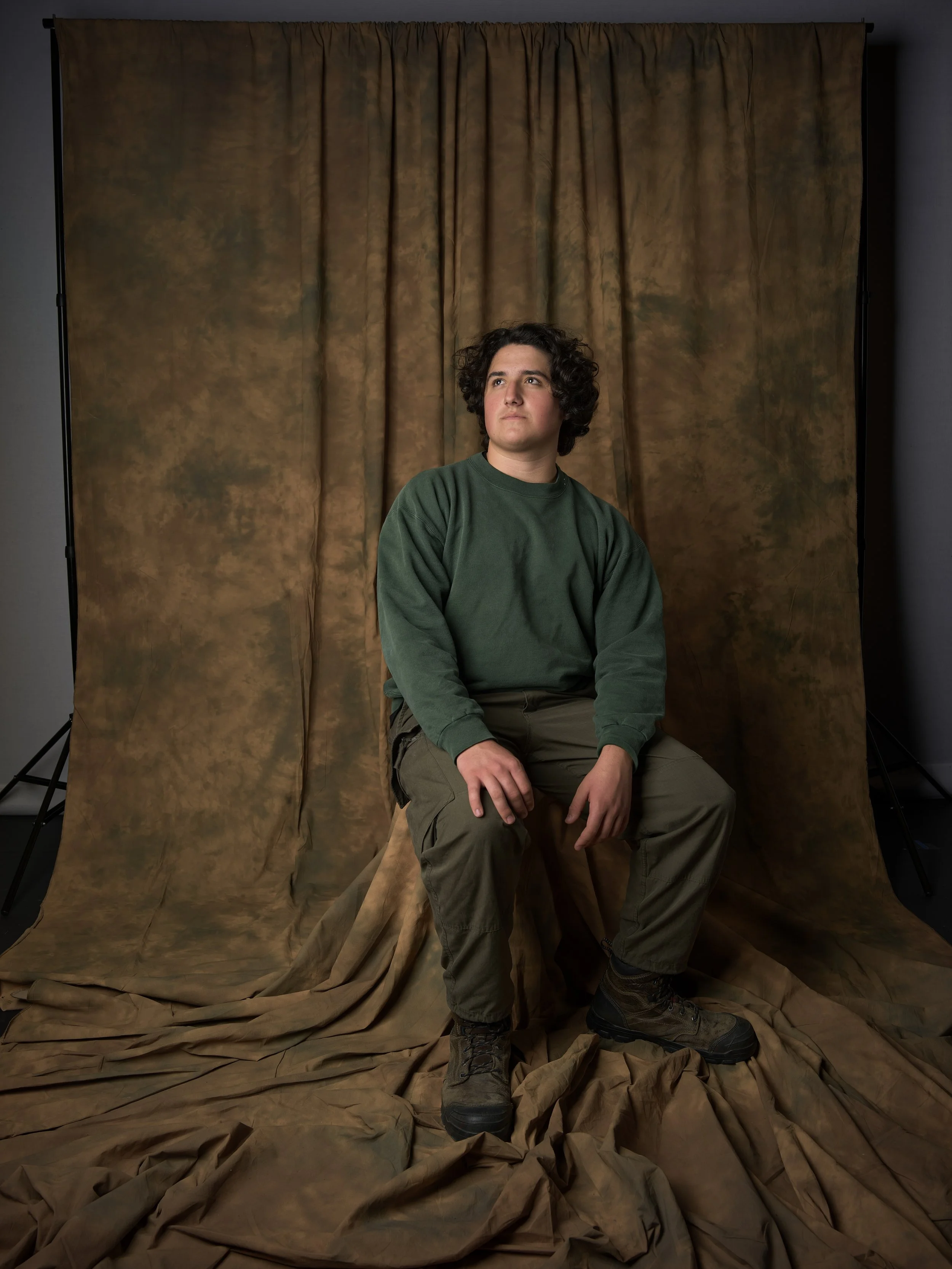 A young man with curly dark hair, wearing a green sweatshirt and khaki pants, sitting on a stool in front of a brown backdrop with fabric on the floor.