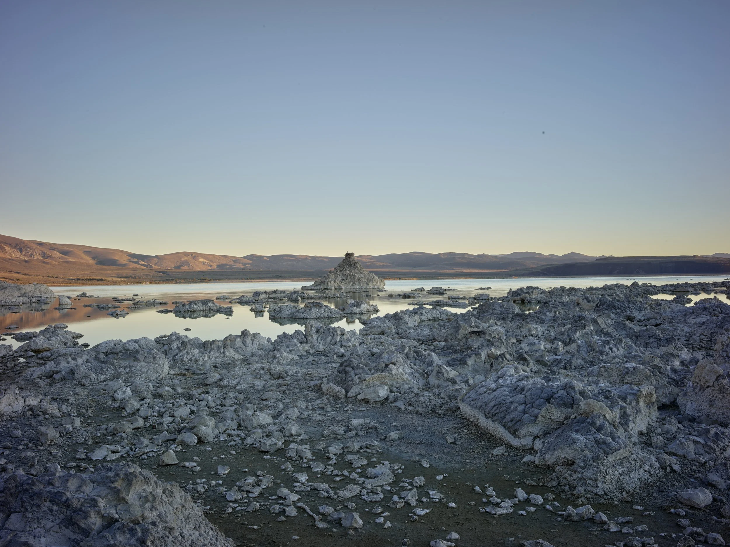 A rocky shoreline with a calm body of water and a small pyramidal formation in the middle, with distant rolling hills and a clear sky in the background.