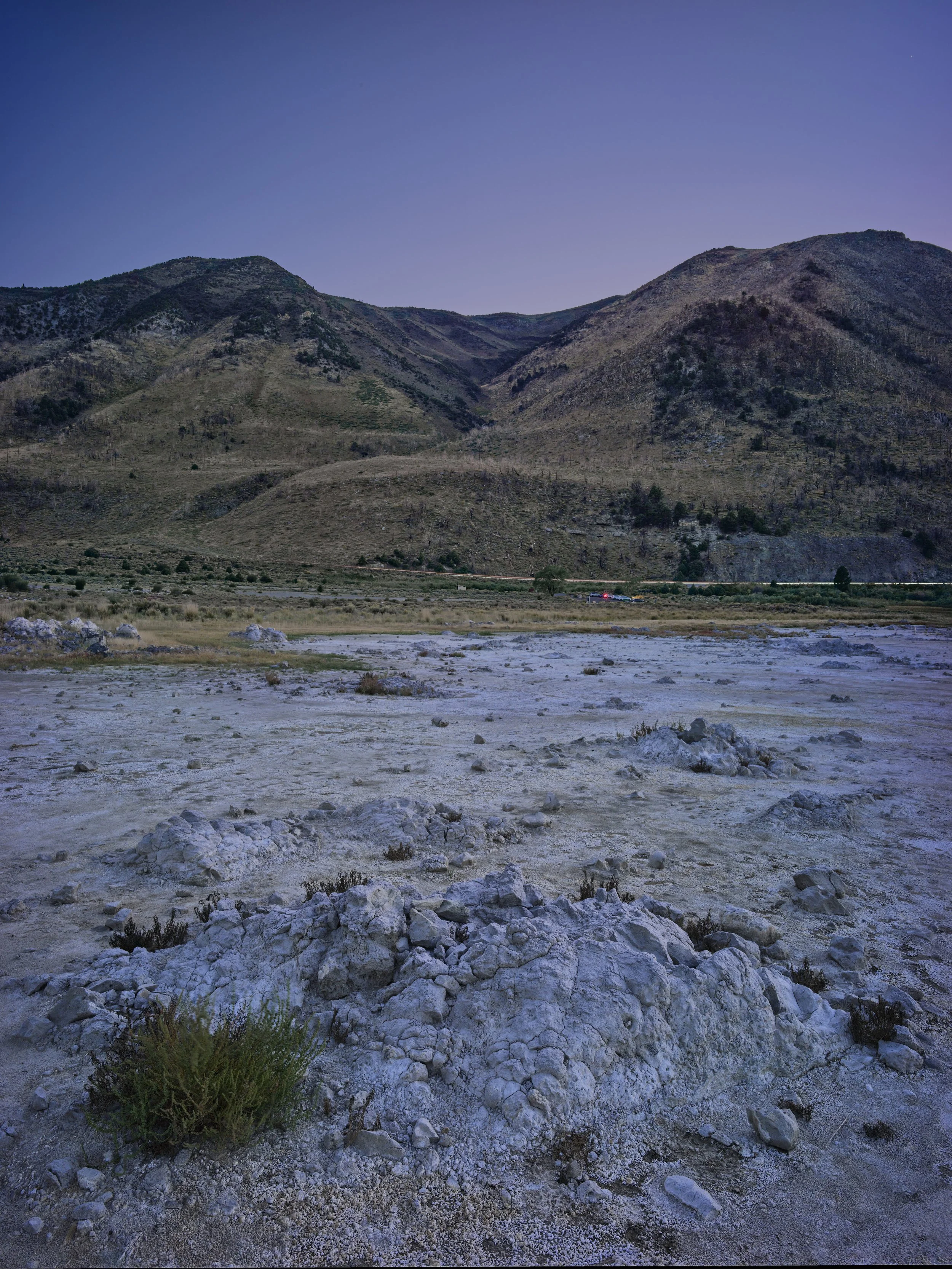 Dry, rocky desert landscape with mountains in the background under a clear evening sky.