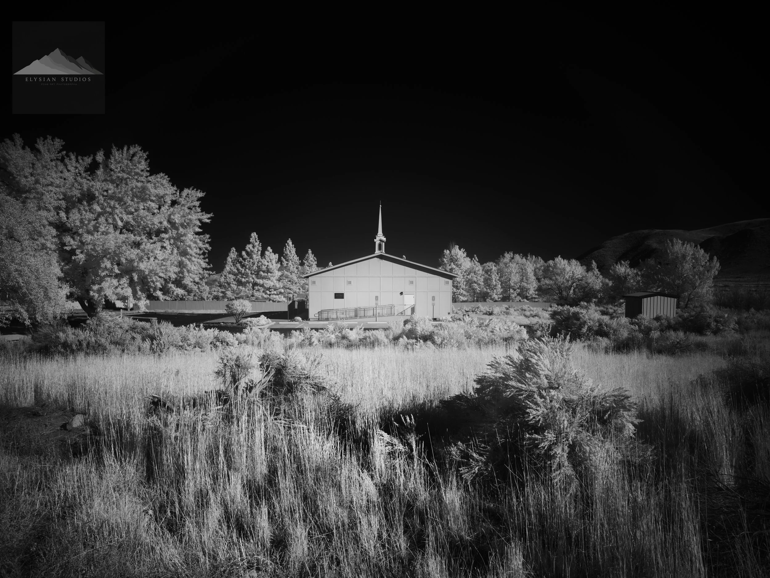 Nighttime scene in black and white featuring a building with a steeple, surrounded by trees and open grassy land with hills in the background.
