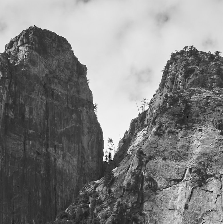 Black and white photograph of tall, rugged mountain cliffs with sparse trees.