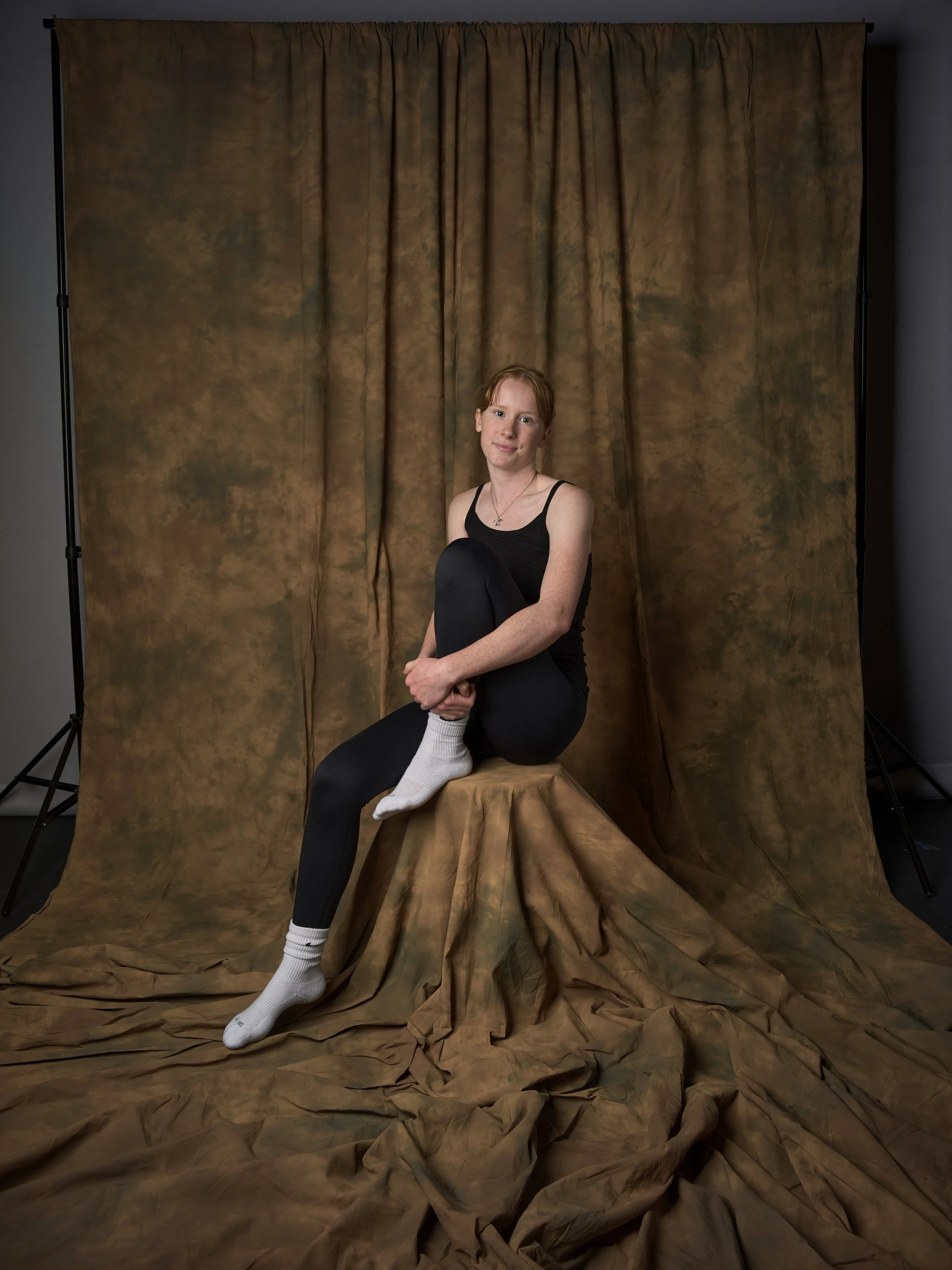 A young woman with red hair sitting on a tree stump, with one leg crossed over the other, in front of a brown fabric backdrop. She is wearing a black tank top, black leggings, and white socks, and is smiling at the camera.