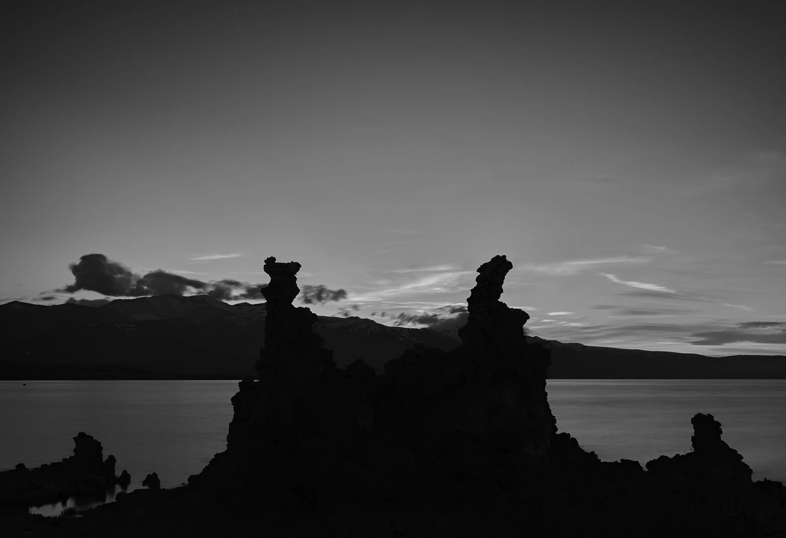Silhouetted rock formations by a lake at sunset or dawn, with mountains and clouds in the distance, in black and white.