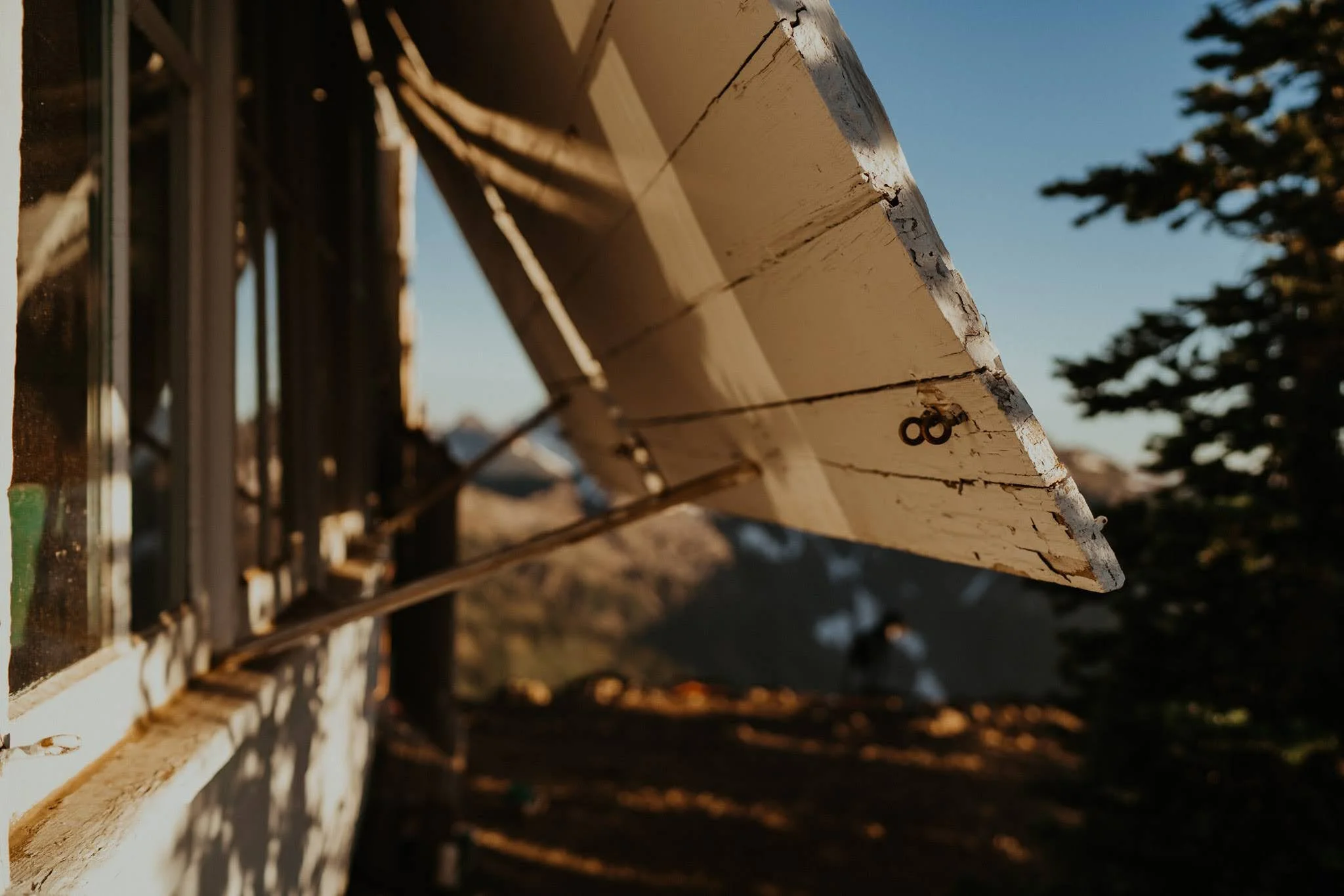 Close-up of a weathered, white wooden airplane propeller mounted on a building, with a blurred background of trees and mountains during sunset.