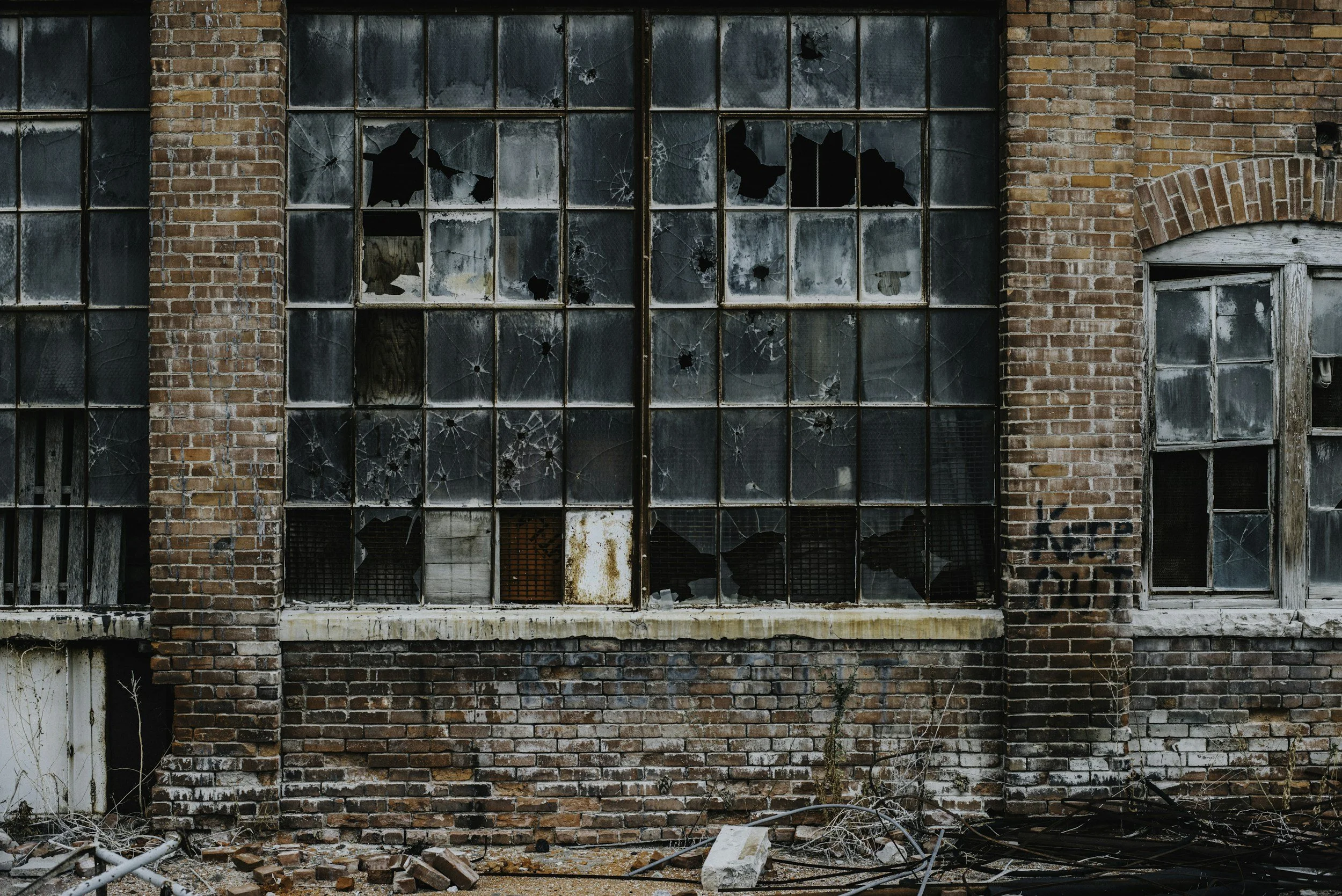 Broken and shattered windows on an old brick building, with some windows boarded up, debris on the ground, and graffiti on the wall.