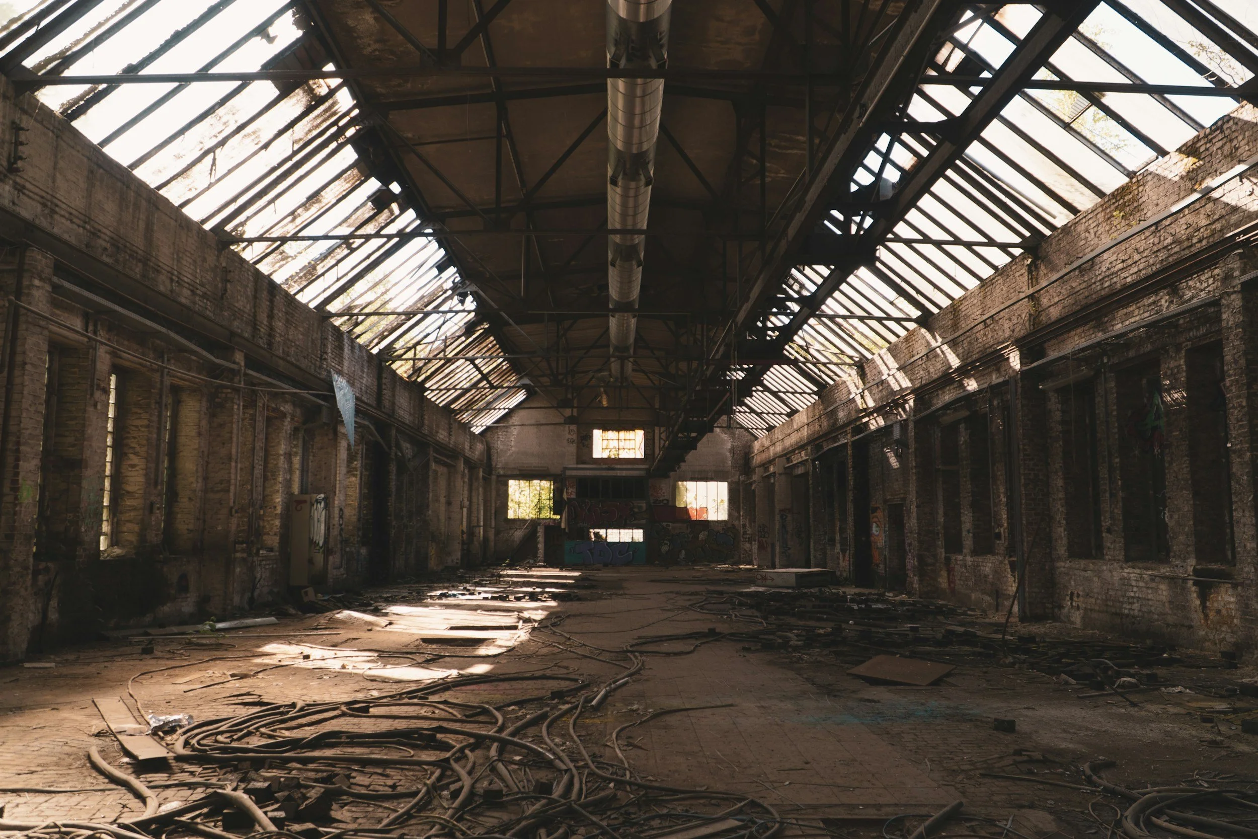 An abandoned industrial building with broken windows, debris on the floor, and graffiti on the walls. Natural light filters through the broken roof.