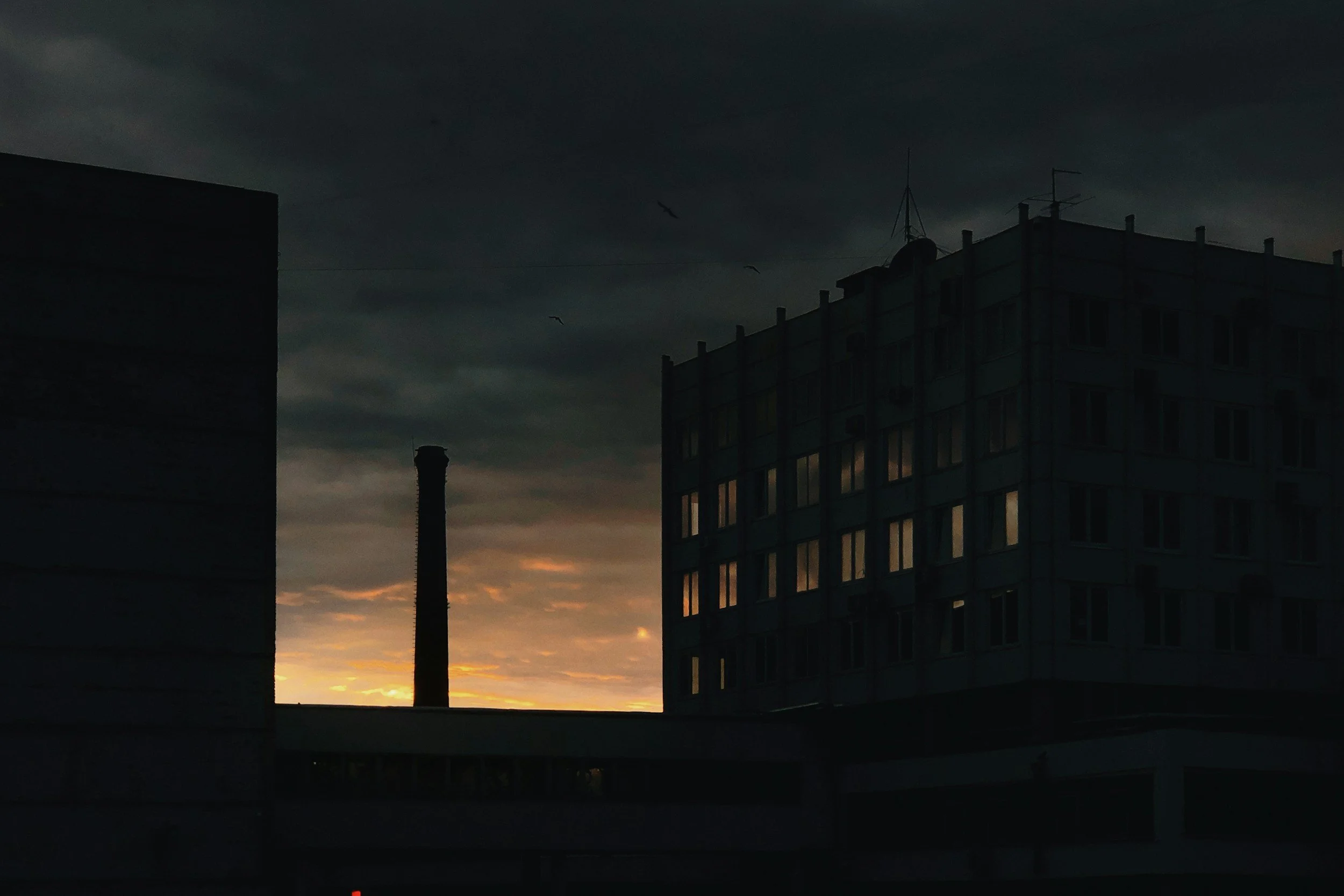 Silhouetted buildings against a dark, cloudy sky at sunset with a few birds flying.