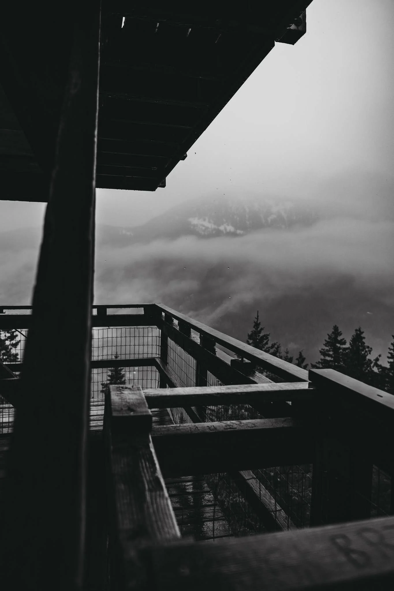 Black and white photo of a wooden observation deck with railings, overlooking foggy mountains and trees in the distance.