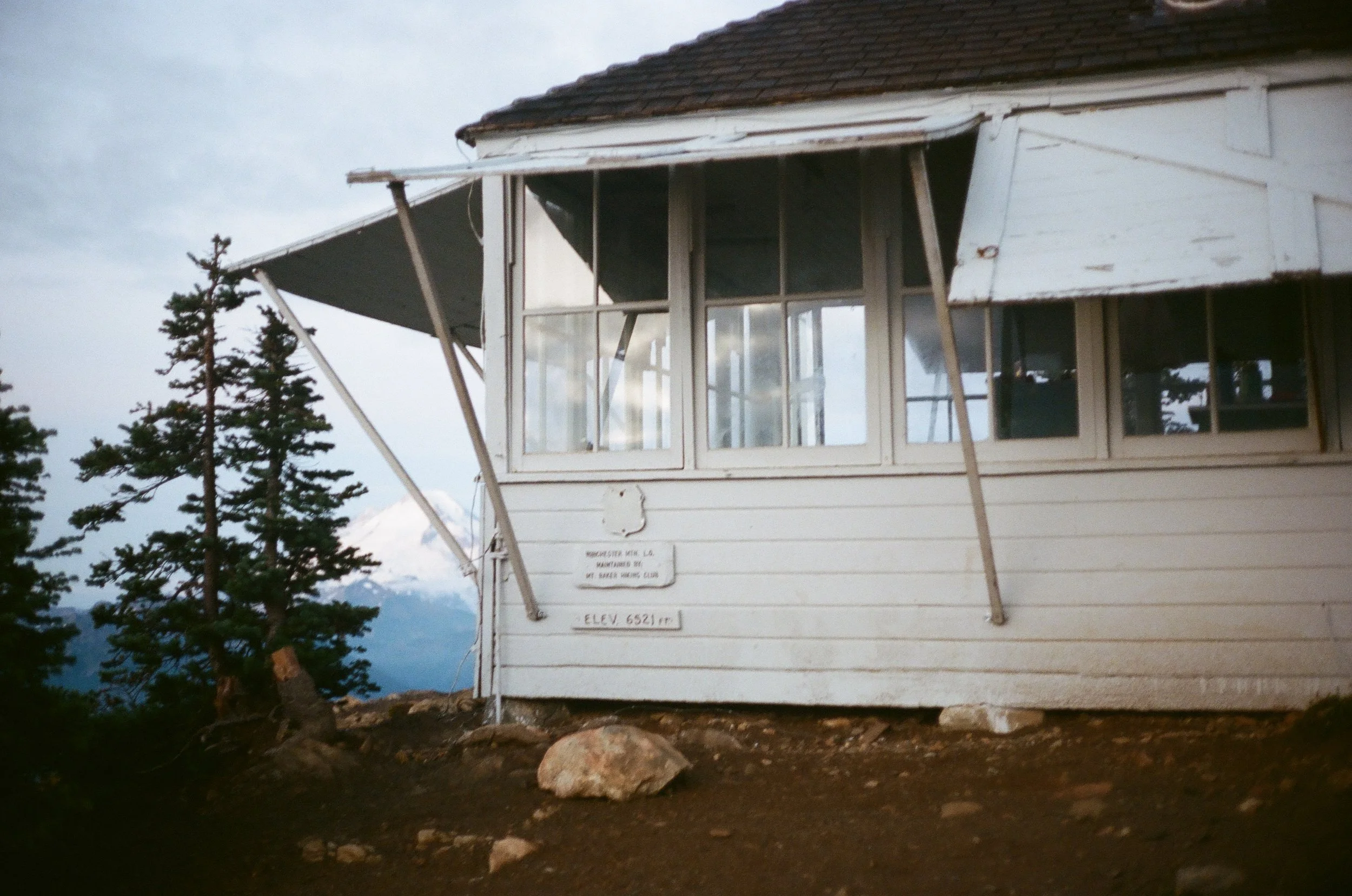 A white mountain cabin with broken windows, supported by metal braces, on a dirt lot with rocks and trees, with a snowy mountain in the background.