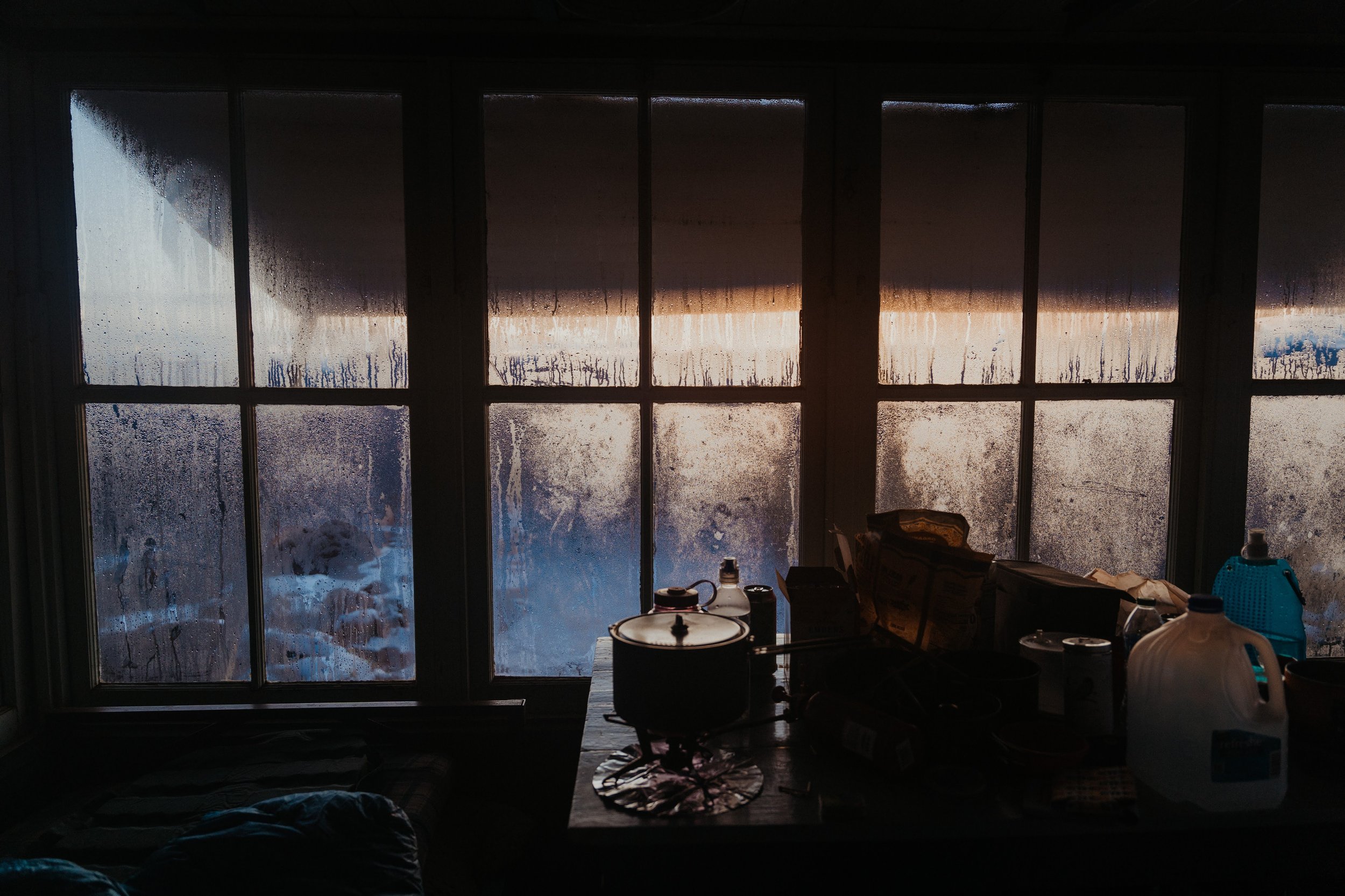 A dark kitchen with a large window fogged up from condensation, showing an outdoor scene of snow and trees at sunset. The kitchen counter has various items including pots, bottles, and boxes.
