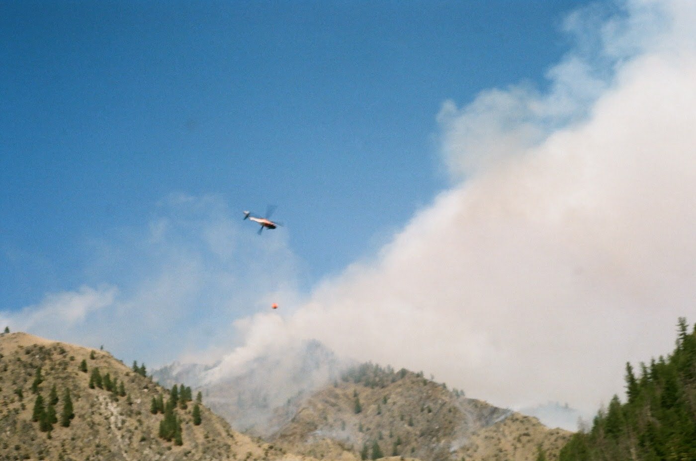 A helicopter flying near a mountain with smoke and clouds in the background.
